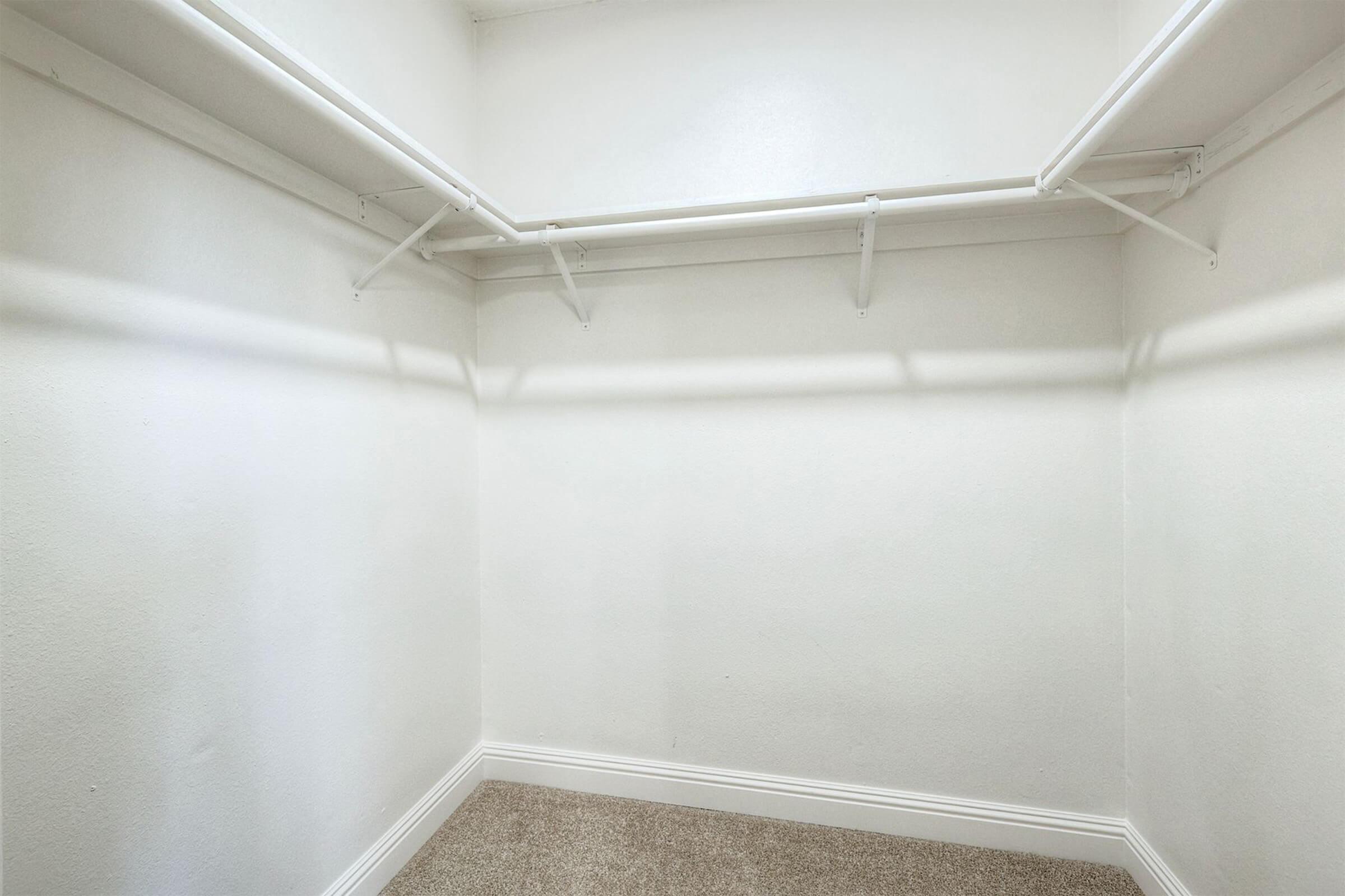 Empty closet with white walls, a light-colored carpeted floor, and no visible items or shelves except for two hanging rod brackets mounted on the walls. The space is well-lit and has a minimalistic appearance, emphasizing its emptiness.