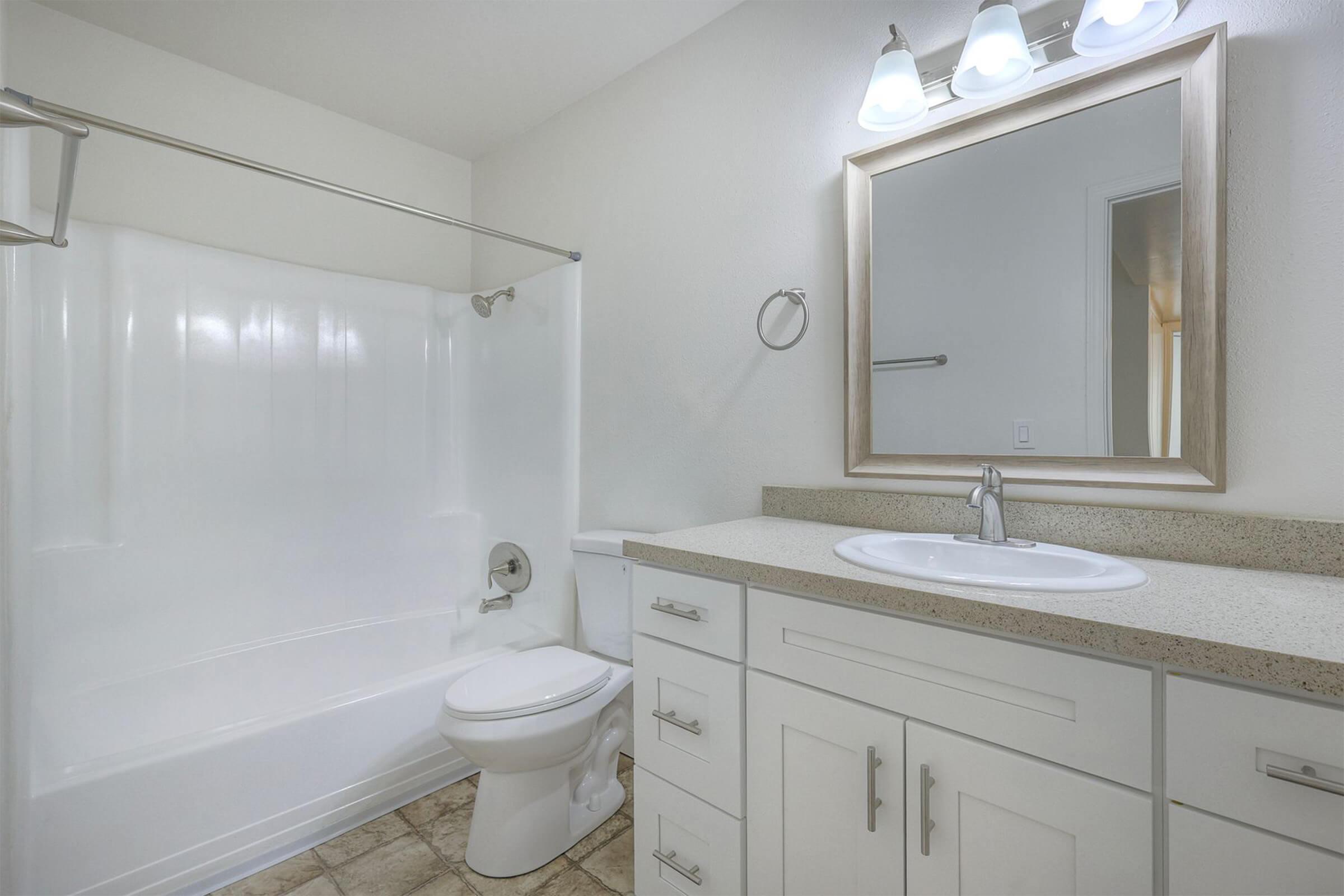 A clean, modern bathroom featuring a white bathtub with shower, a toilet, and a light-colored countertop sink. The room has neutral-toned walls and a large mirror above the sink, illuminated by three wall-mounted light fixtures. The flooring is a textured tile.