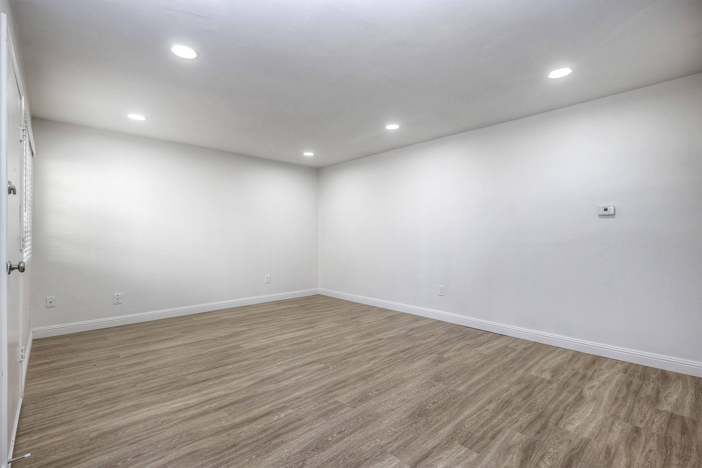 A spacious, empty room with smooth white walls and a light-colored wooden floor. Soft, recessed ceiling lights provide bright illumination. There are no furniture or decorations, creating a minimalist and open atmosphere. A thermostat is visible on one wall, adding functional detail to the otherwise bare space.