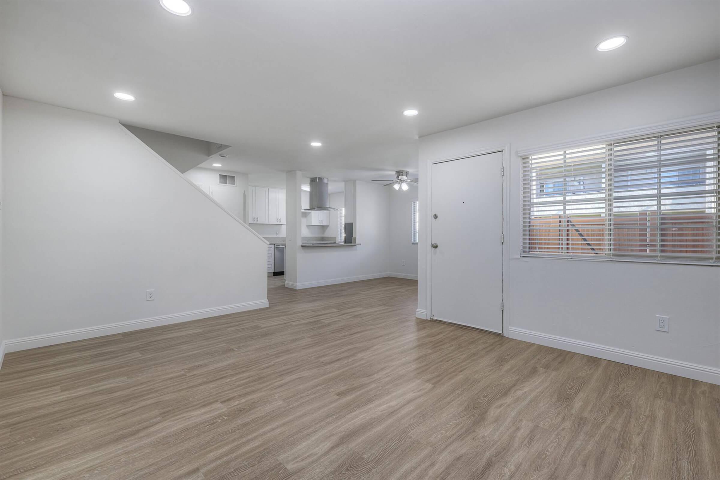 Bright, open living space with light-colored walls and wood-style flooring. A staircase is visible on the left, and a door leads outside. A window with blinds allows natural light, and the kitchen area can be seen in the background with modern white cabinetry. The overall atmosphere is clean and contemporary.
