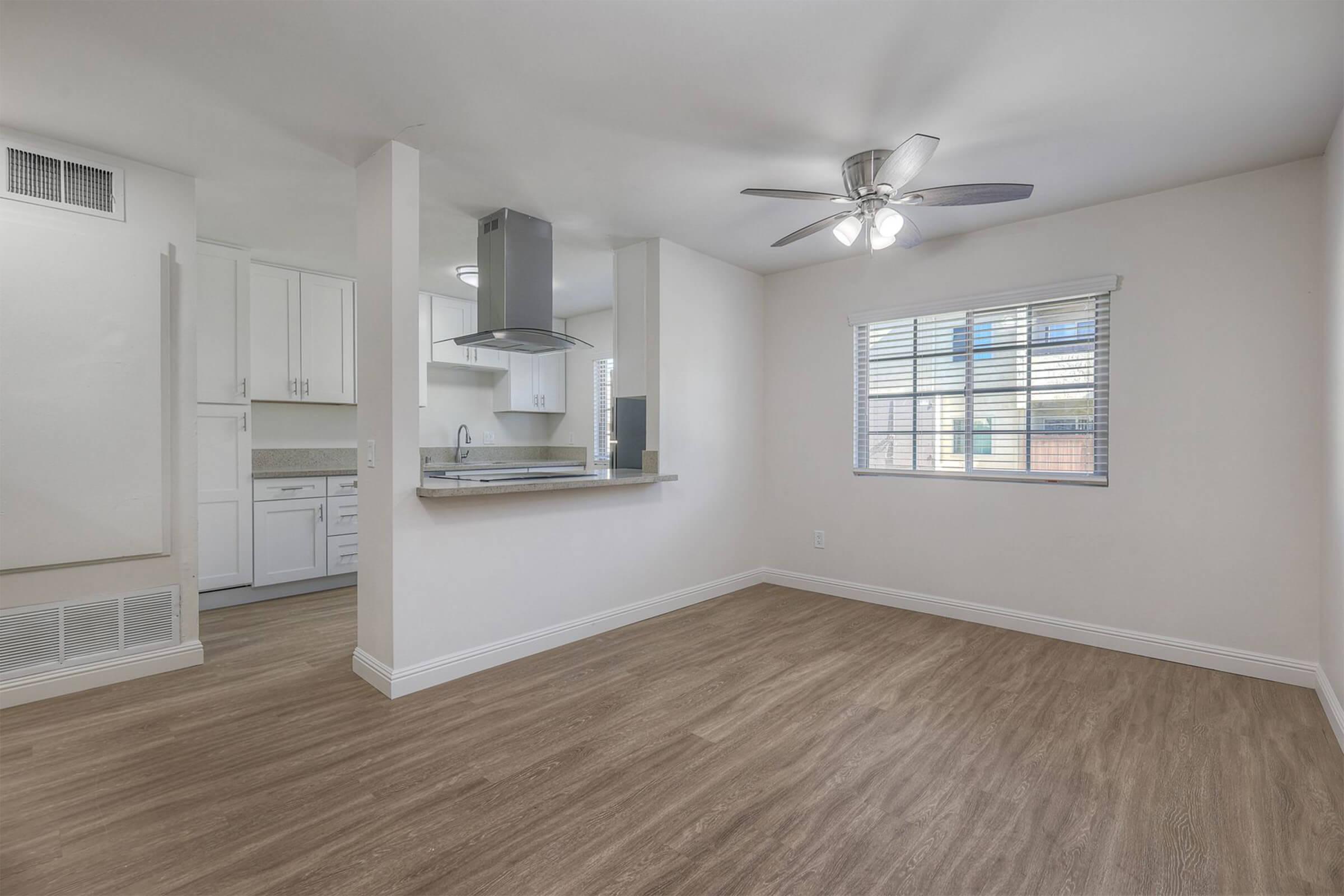 Bright and spacious living area with a ceiling fan, featuring hardwood-style flooring. An open kitchen area with white cabinetry is visible in the background, along with a window that allows natural light to enter the room. The overall atmosphere is modern and inviting.
