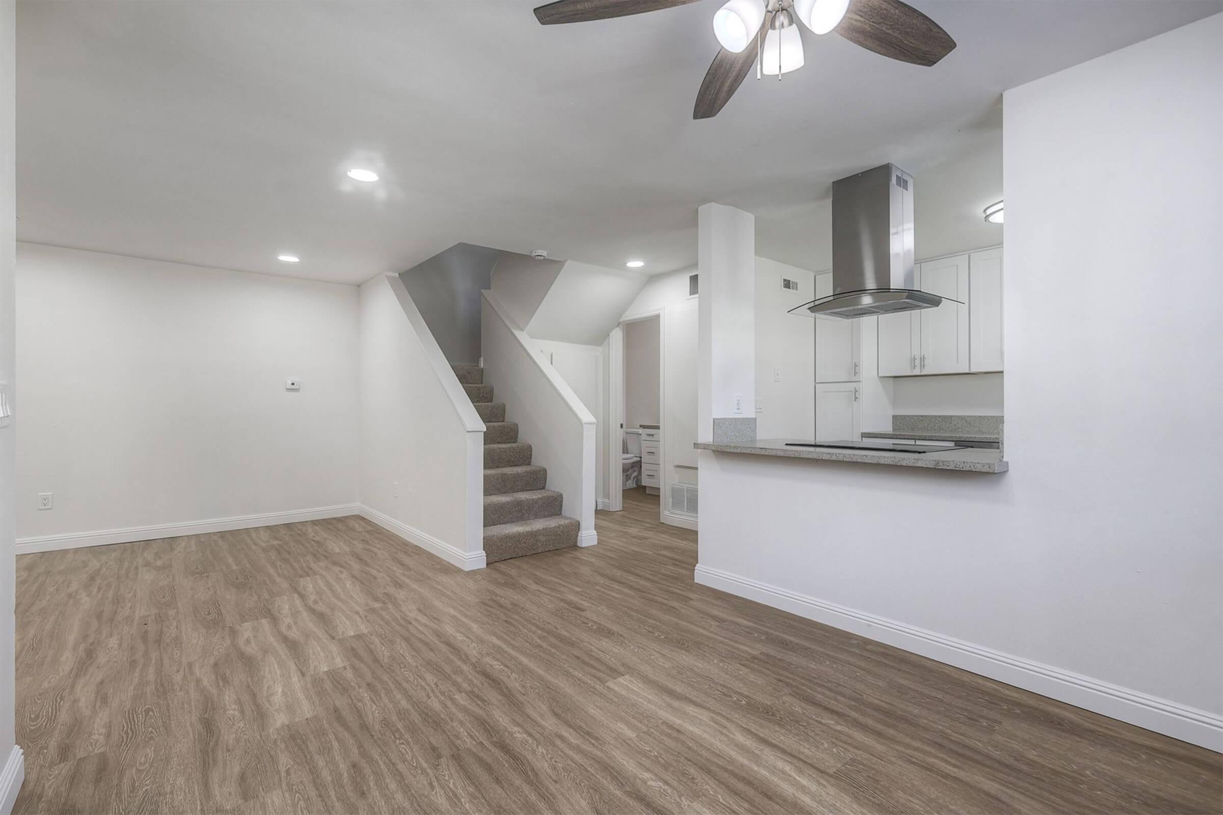 Spacious living area featuring wood-style flooring, a ceiling fan, and a staircase leading to the upper level. The open layout connects to a modern kitchen with white cabinetry. Natural light enters through the windows, creating a bright and welcoming atmosphere.