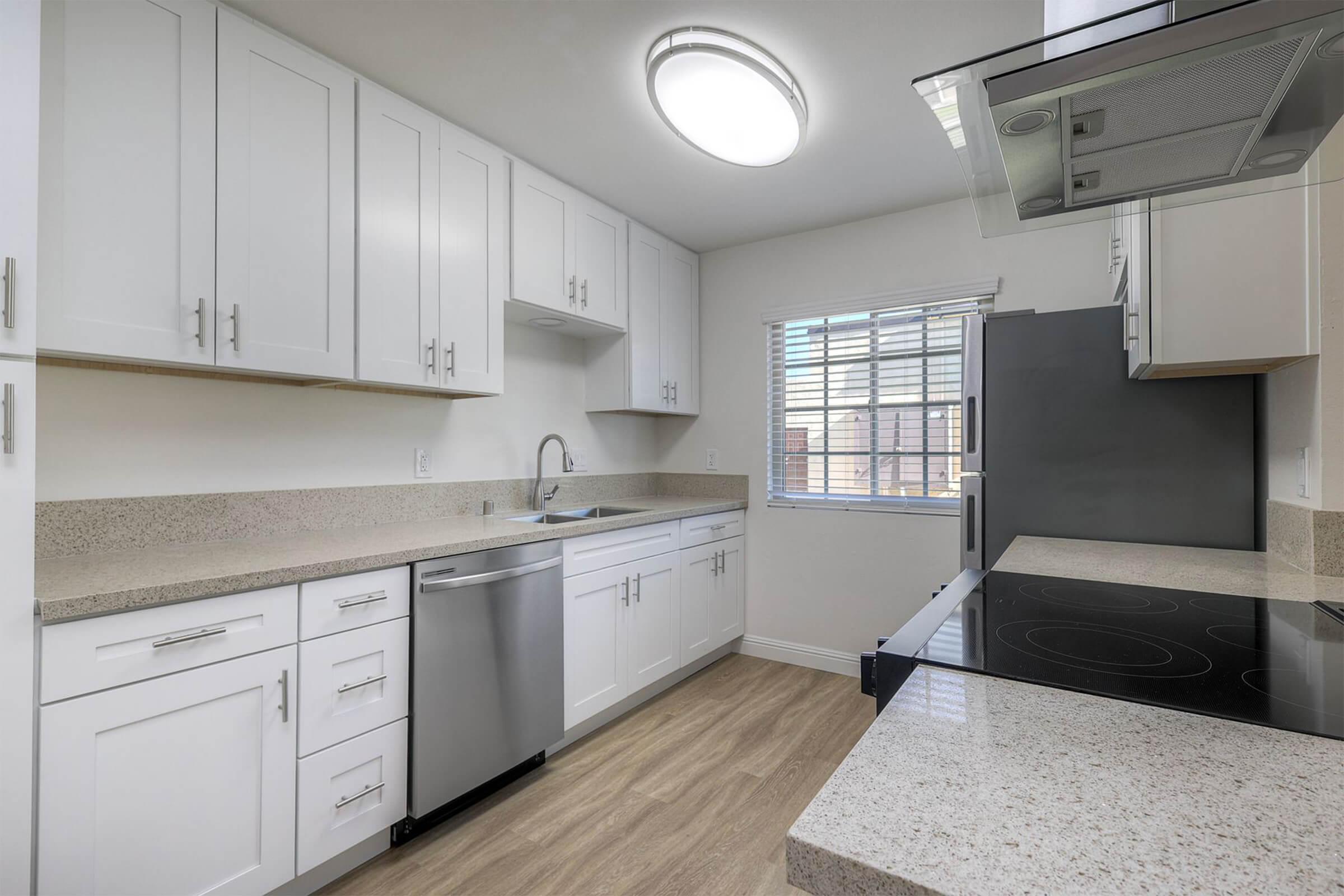 Modern kitchen featuring white cabinetry, stainless steel appliances, and a sleek countertop. The space includes a sink, a dishwasher, and an electric cooktop, with a window that provides natural light. Flooring is light wood laminate, creating a bright and airy atmosphere.