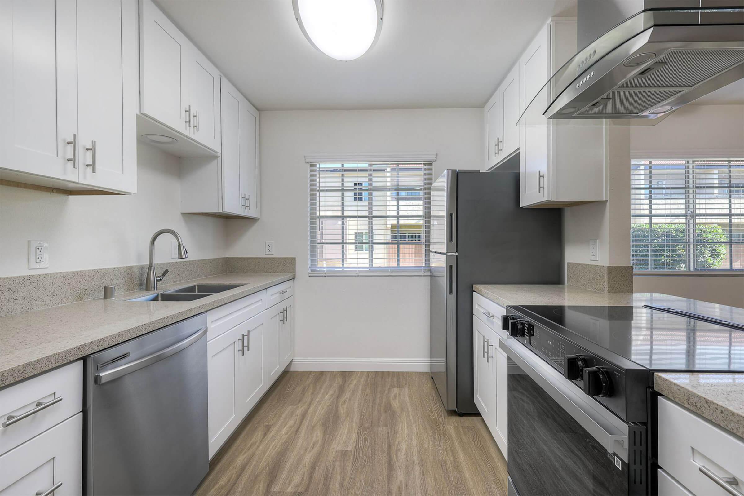 Modern kitchen featuring white cabinetry, stainless steel appliances, and granite countertops. Includes a sink, dishwasher, refrigerator, and stove, with a window allowing natural light and wood-look flooring. Bright and functional layout ideal for cooking and meal preparation.