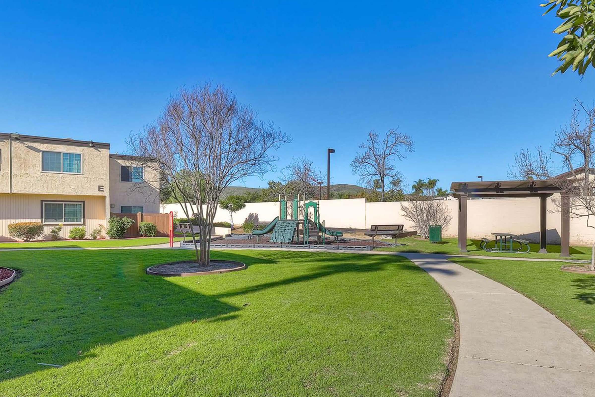 A sunny outdoor area featuring a grassy lawn, playground equipment, and benches. In the background, there are two residential buildings with a clear blue sky above. Leafless trees add to the scenery, creating a welcoming environment for families and children.