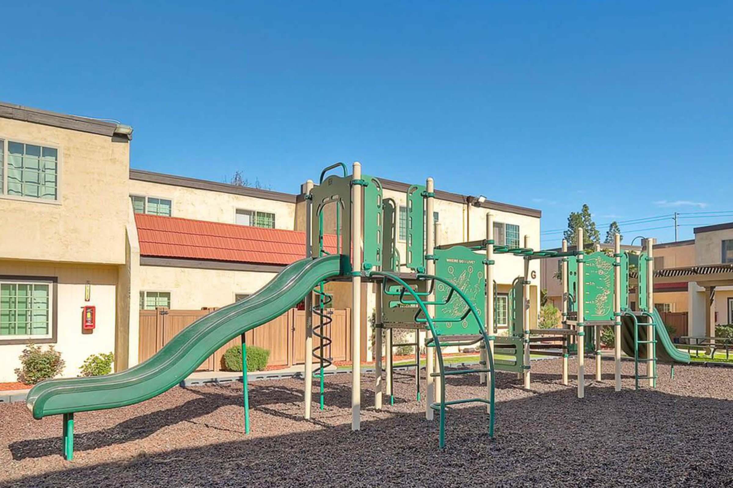 Playground equipment featuring a slide and climbing structures, set in a gravel area with a clear blue sky. In the background, there are buildings with green windows and a red roof. The scene is inviting and designed for children to play and explore.