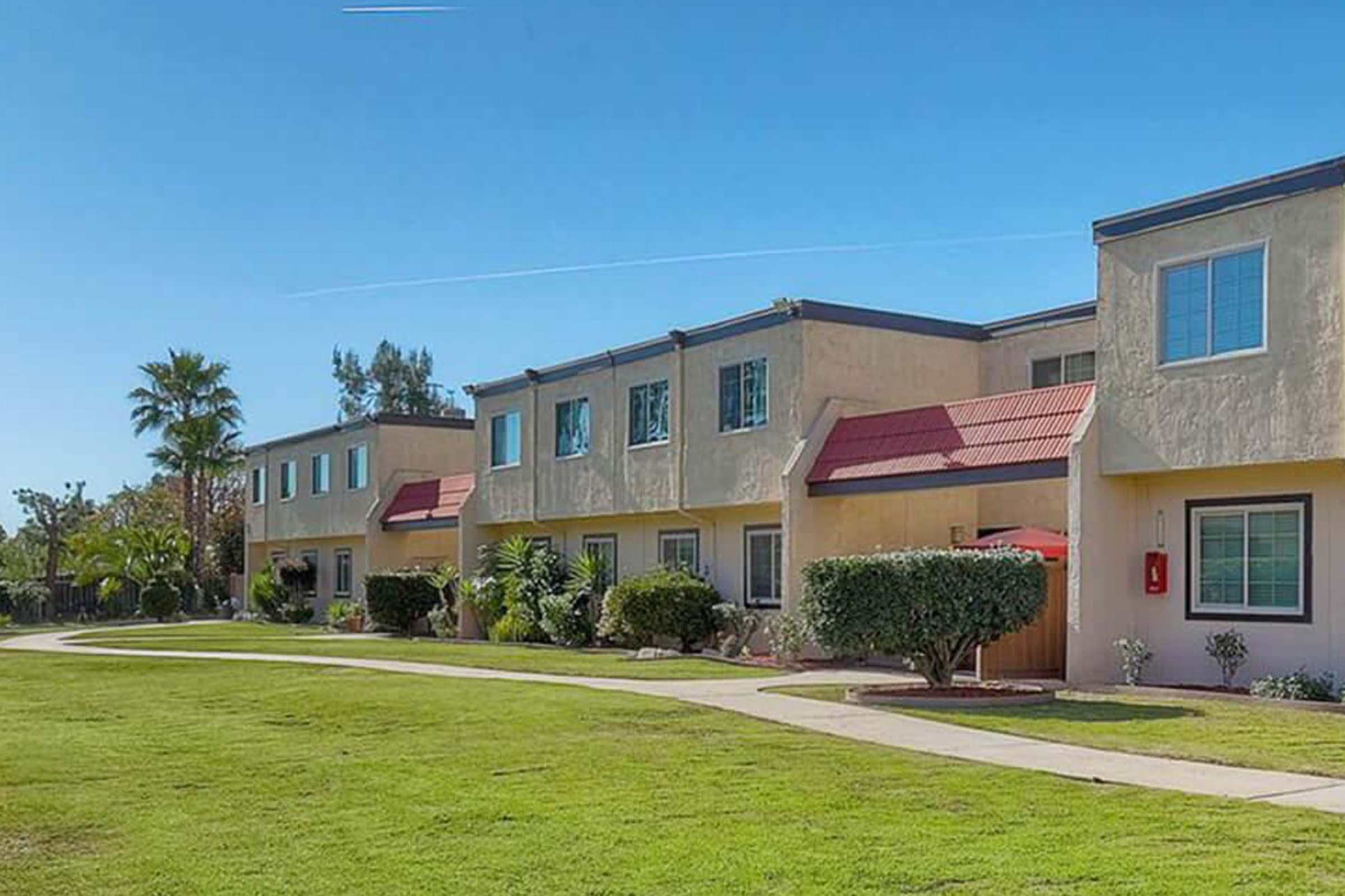 A row of townhouses with cream-colored exteriors and red roofs, set against a clear blue sky. The landscape features well-maintained green grass and small shrubs, along with palm trees, creating a pleasant and inviting residential atmosphere.