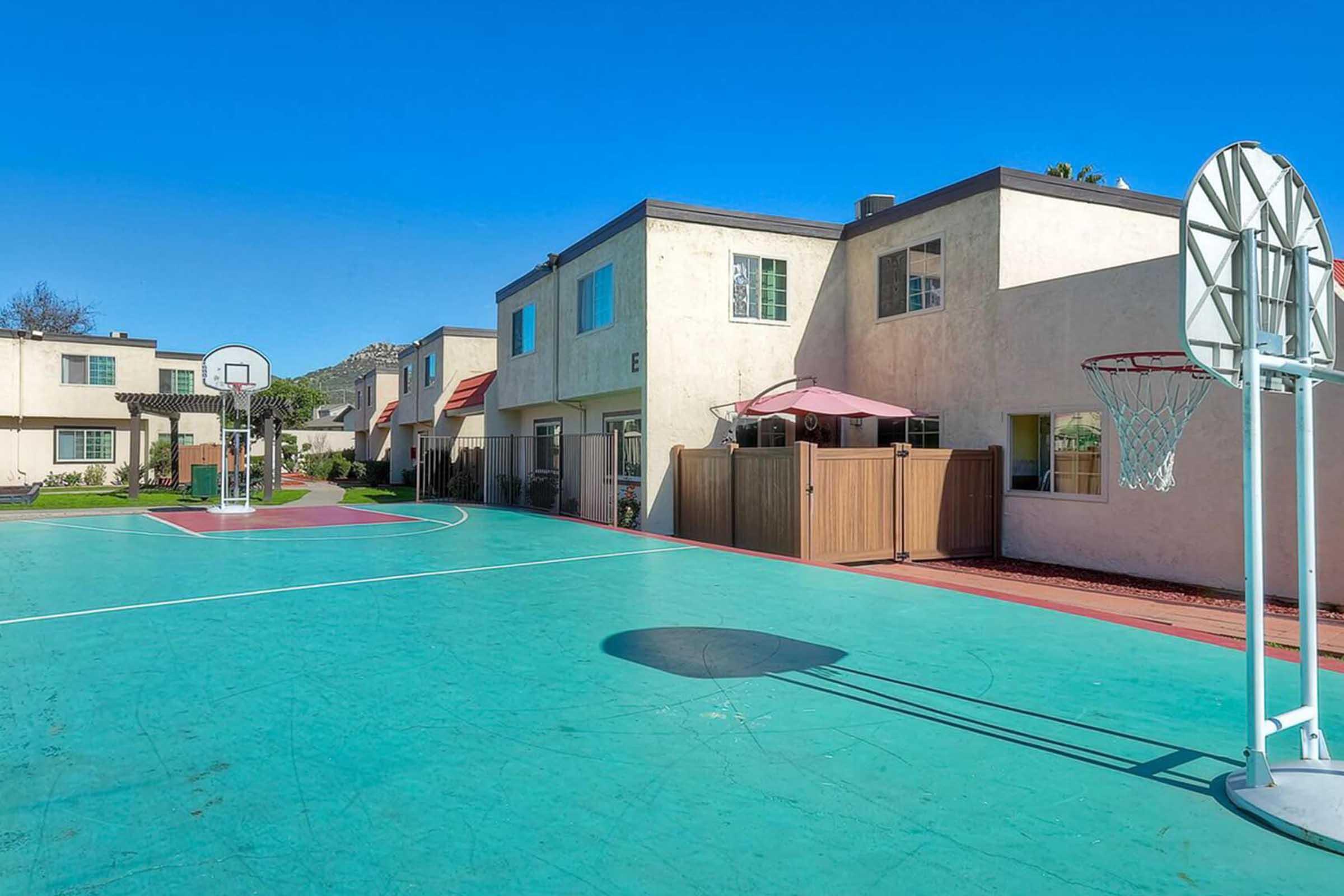A basketball court featuring a hoop on the right side, surrounded by residential buildings. The court's surface is green with red accents, and there's a shaded seating area with an umbrella nearby. The sky is clear and blue, creating a bright atmosphere.