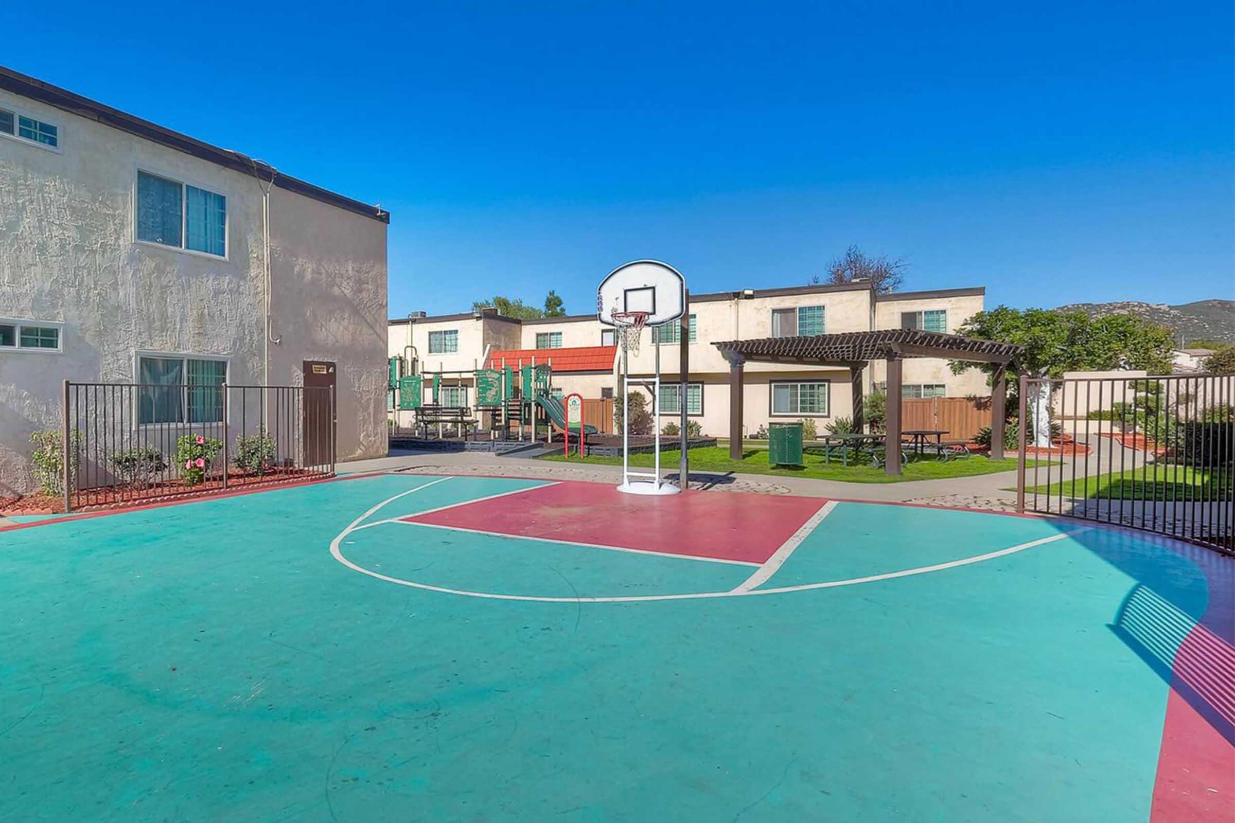 A basketball court with a hoop in the foreground, surrounded by colorful playground equipment and residential buildings. The court has distinct green and red markings on the surface, and the sky is clear and blue. Lush greenery and a shaded area can be seen in the background.