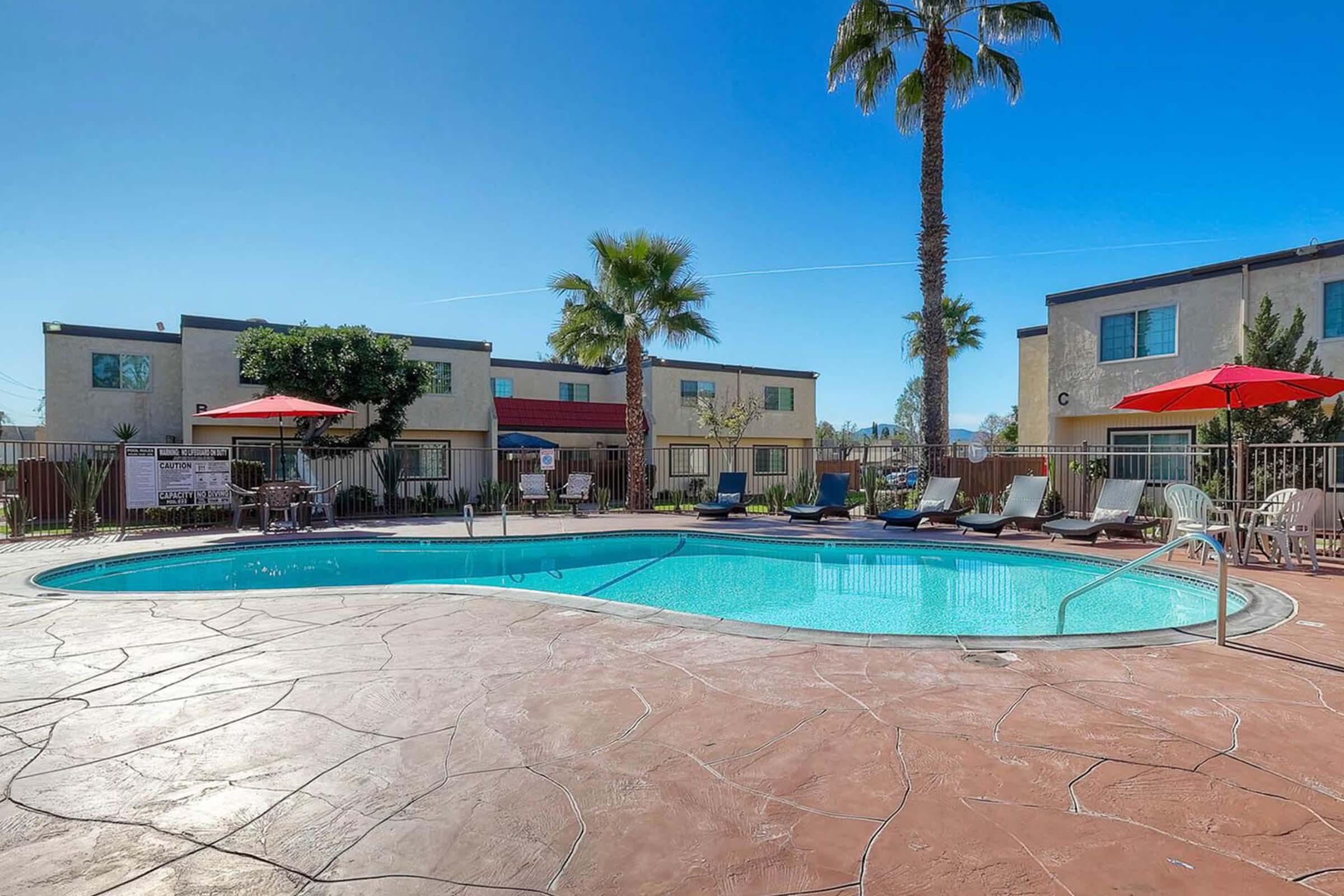 A sunny outdoor pool area featuring a clear blue pool surrounded by lounge chairs, palm trees, and red umbrellas. In the background, there are multi-story buildings, creating a relaxing atmosphere for guests.