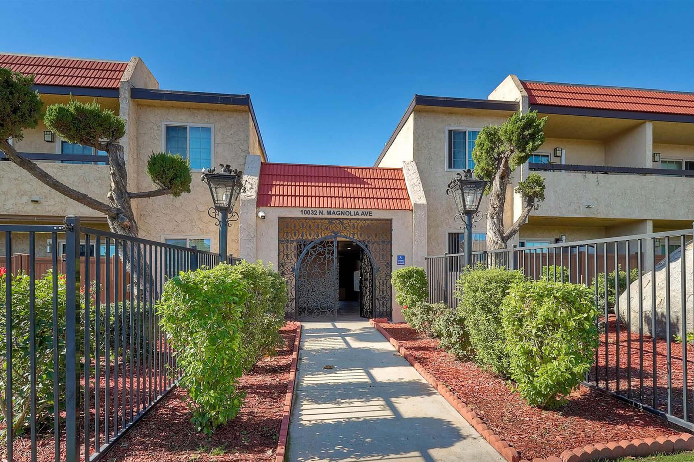 Entrance to an apartment complex featuring a decorative gate, flanked by landscaped shrubs and small trees. The building has a beige exterior with a red tile roof, and two stories are visible under a clear blue sky. The pathway leads to a central doorway, adding to the welcoming atmosphere.