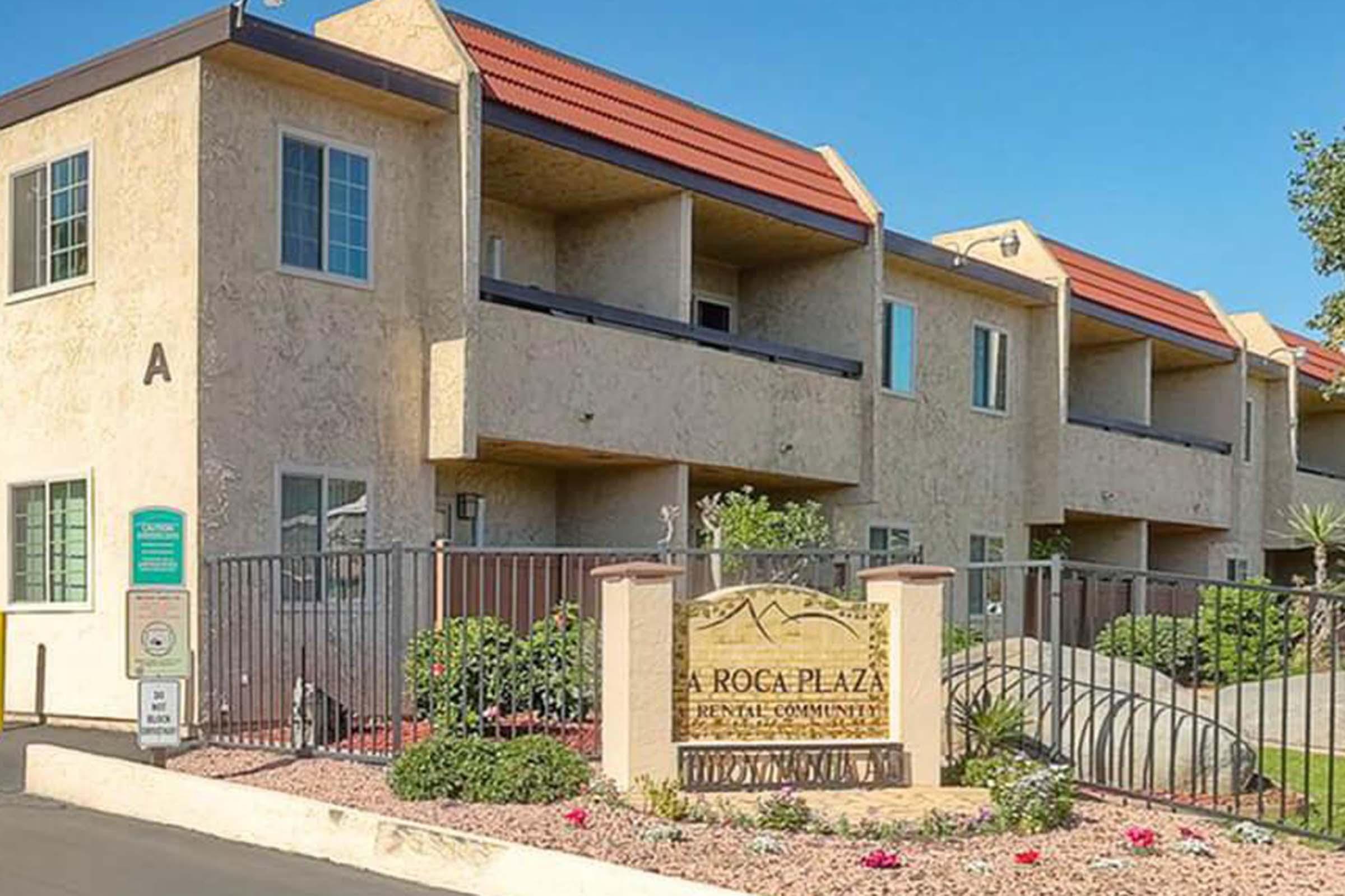 Exterior view of Roca Plaza, a rental community. The building features a beige exterior with red roofs, a well-maintained garden, and a decorative sign at the entrance. A gated area and nearby landscaping are visible, showcasing a pleasant residential environment.