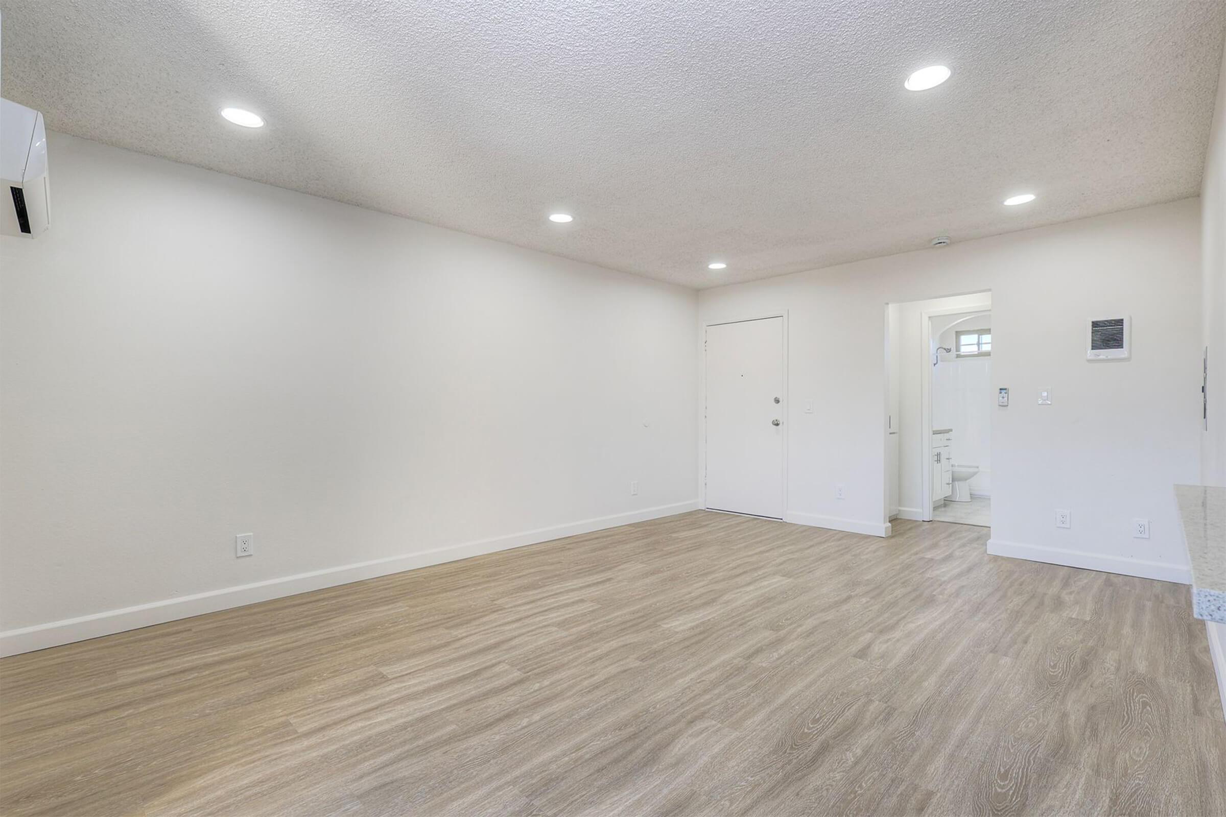 Empty room with light-colored walls and a textured ceiling. The floor is finished in light wood laminate. A wall-mounted air conditioning unit is on the left, and a white door leads to a bathroom visible in the background. The space is well-lit with recessed lighting.