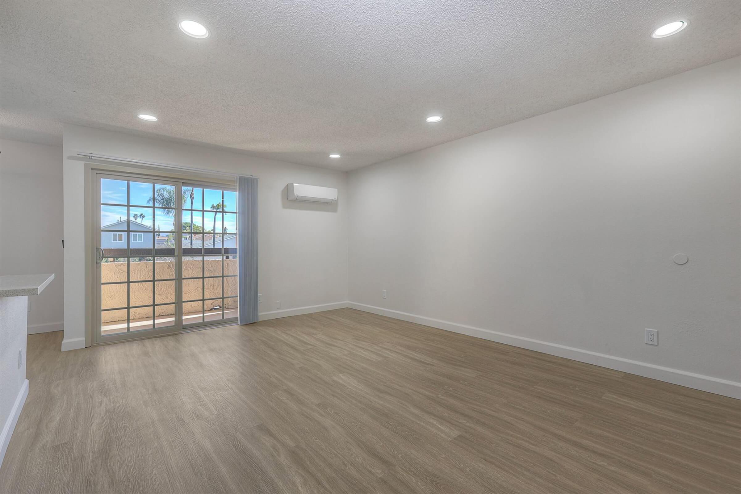 A spacious, unfurnished living room featuring light-colored walls, a large sliding glass door with a view of outdoor palm trees, and modern light fixtures. The floor is covered with light wood-like laminate, creating a bright and airy atmosphere. An air conditioning unit is mounted on the wall.