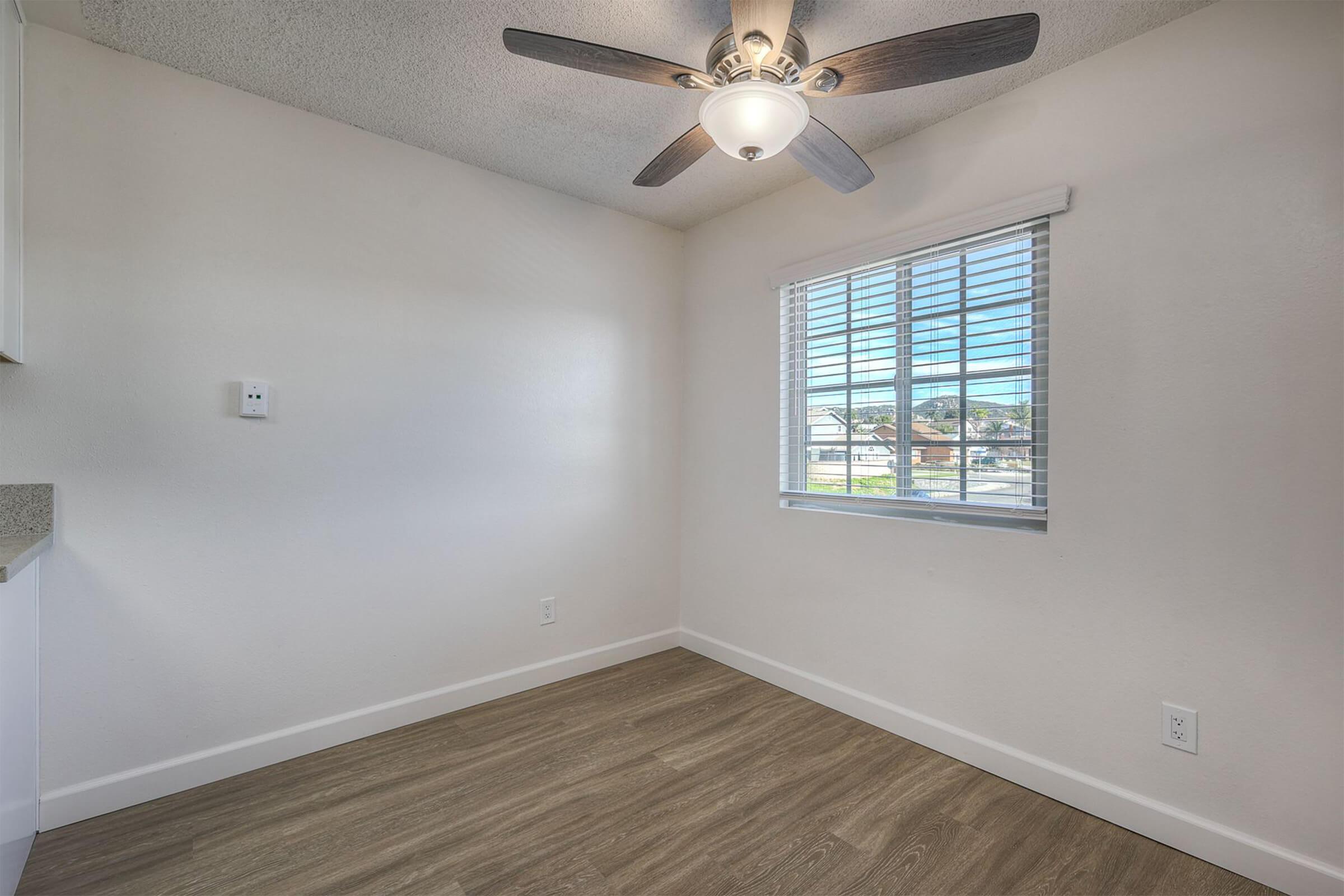 A bright, empty room with light-colored walls and a ceiling fan. There is a window with blinds, allowing natural light to enter. The floor is wooden, and there is a wall-mounted light switch. The space appears clean and ready for furniture.