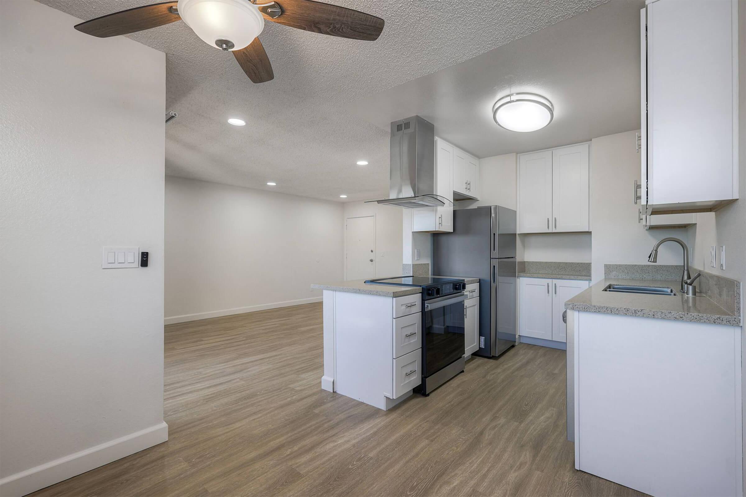 Modern kitchen and living area featuring a ceiling fan, stainless steel appliances, white cabinetry, and light wood flooring. The layout is open, with good natural lighting and a minimalist design.