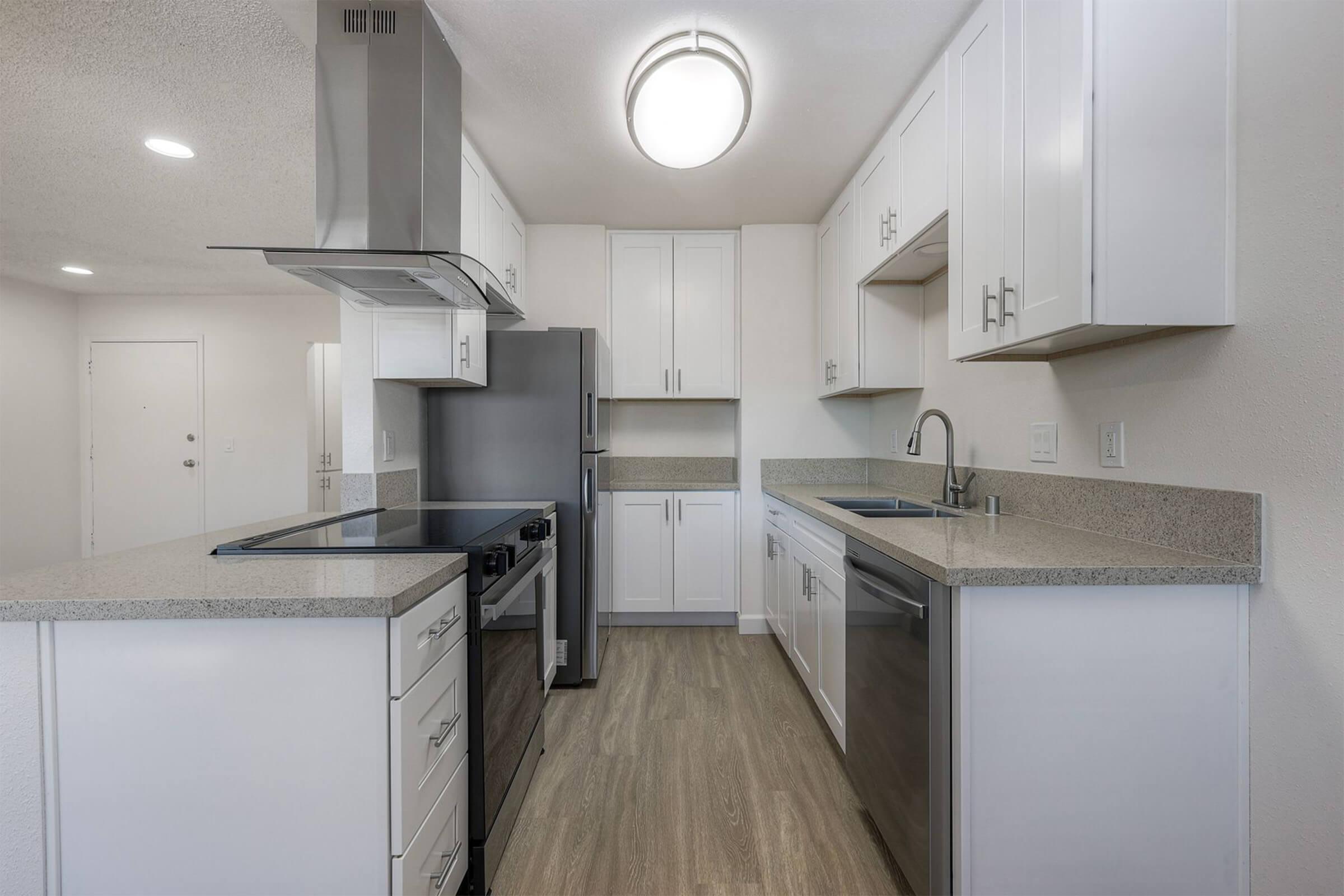 Modern kitchen featuring white cabinetry, a spacious countertop, stainless steel appliances including a refrigerator and stove, a sink with a faucet, and light-colored vinyl flooring. The room is well-lit with a ceiling fixture and has an open layout leading to an adjacent living space.