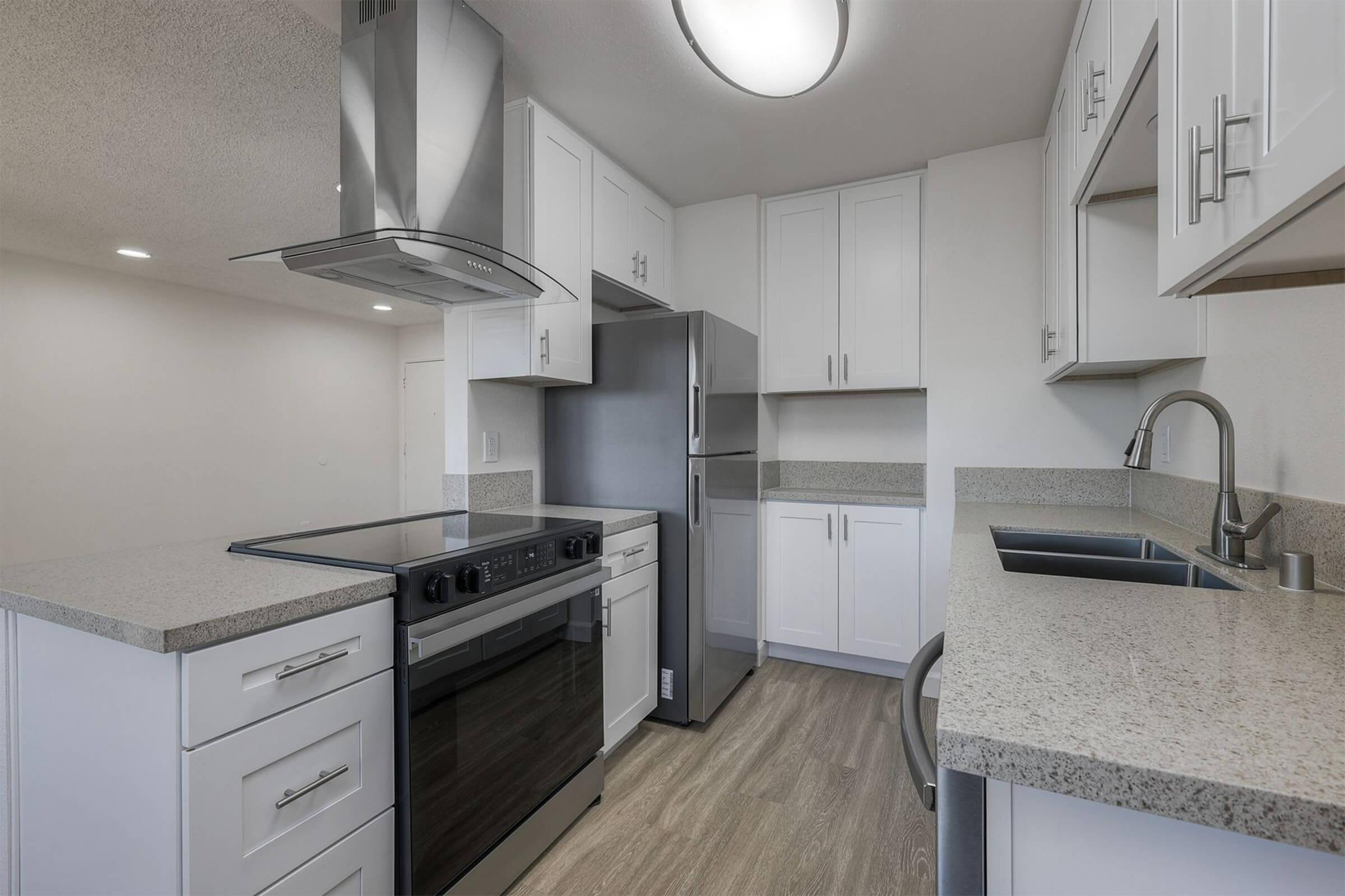 Modern kitchen featuring white cabinetry, a stainless steel refrigerator, an oven, and an overhead range hood. The countertop is light gray with a sink and faucet. The space is well-lit with a ceiling light, showcasing a clean and minimalist design.