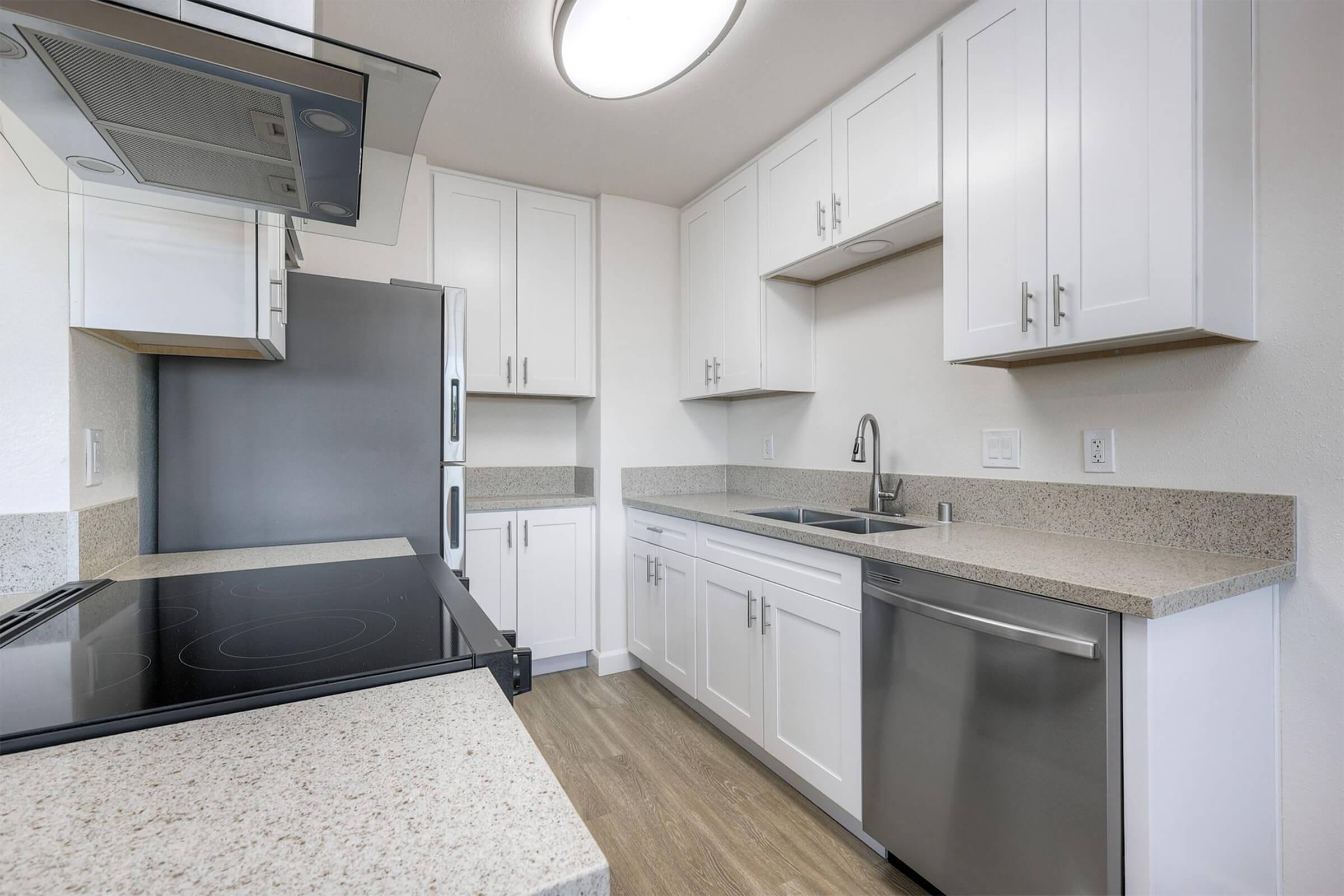 A modern kitchen featuring white cabinets, stainless steel appliances, and a countertop. The space includes a sleek cooktop, built-in oven, refrigerator, and a sink, all arranged in an L-shape with ample natural light from a ceiling fixture.