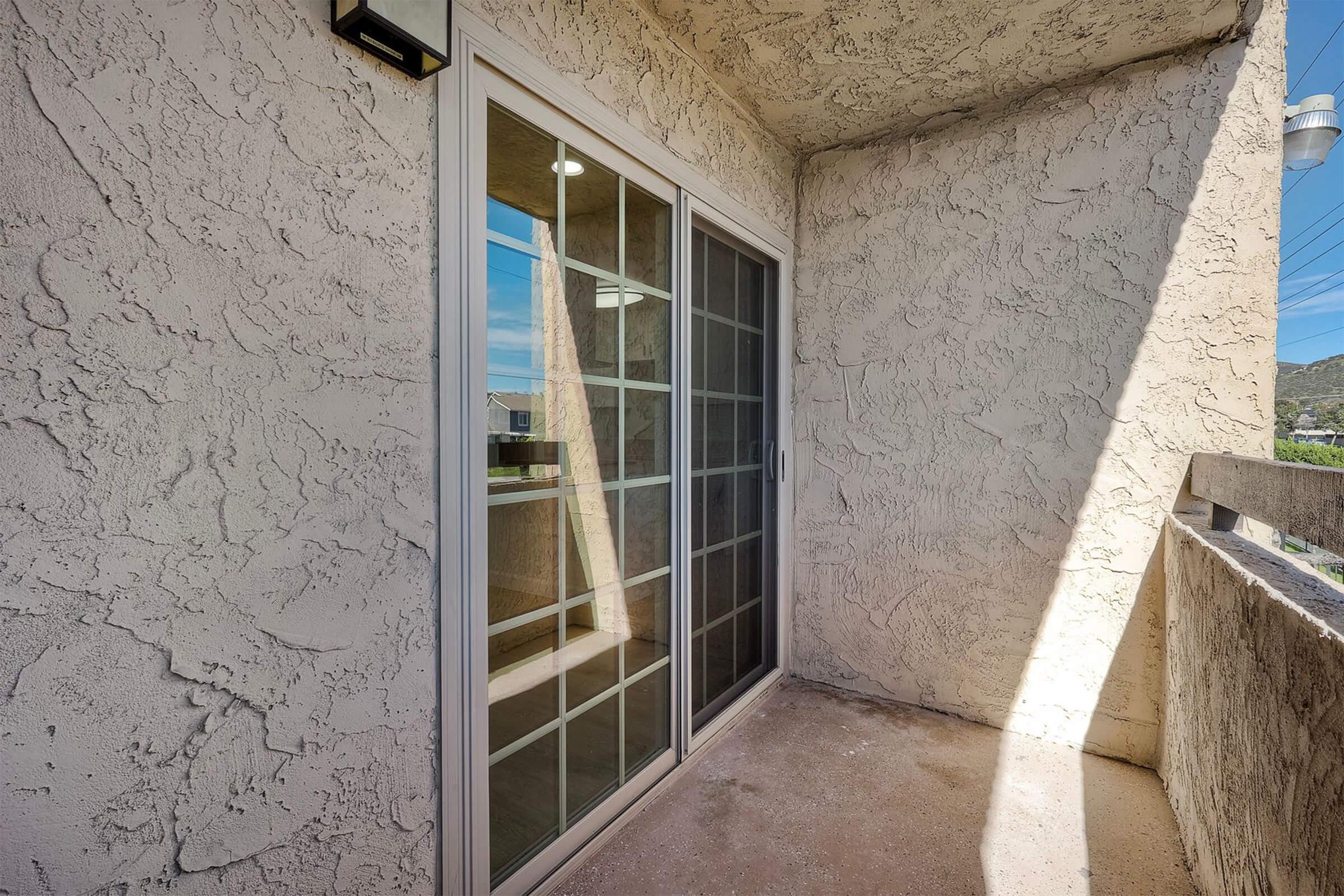A small balcony with a textured beige wall and sliding glass doors, providing access to the outside. The floor is concrete, and the view appears to include a clear sky and distant landscape. Natural light illuminates the space.