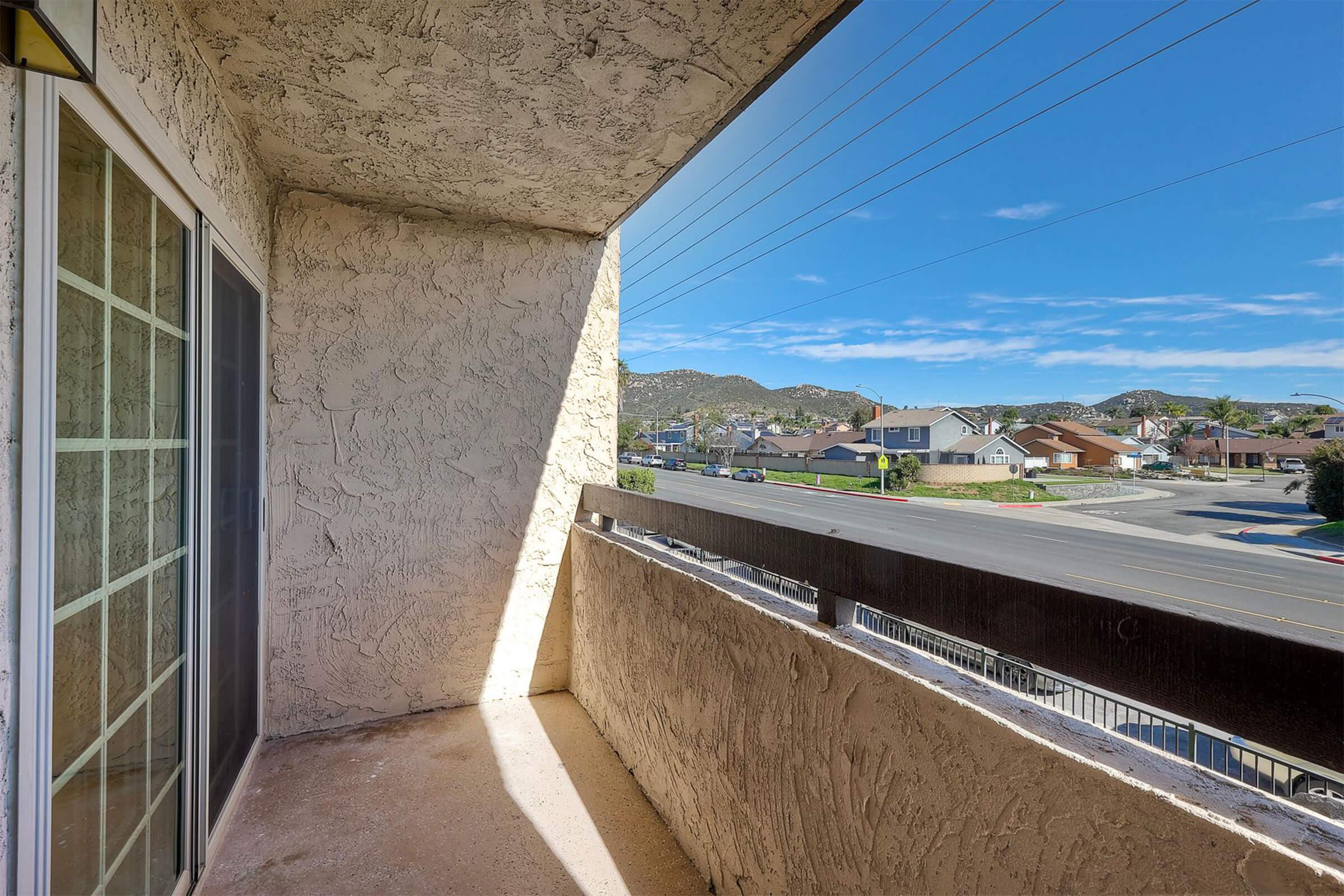 A view from a balcony featuring a concrete surface, looking out over a residential street with houses and mountains in the background. The sky is clear with a few clouds, and utility lines stretch across the image. Sunlight casts a shadow on the balcony floor.