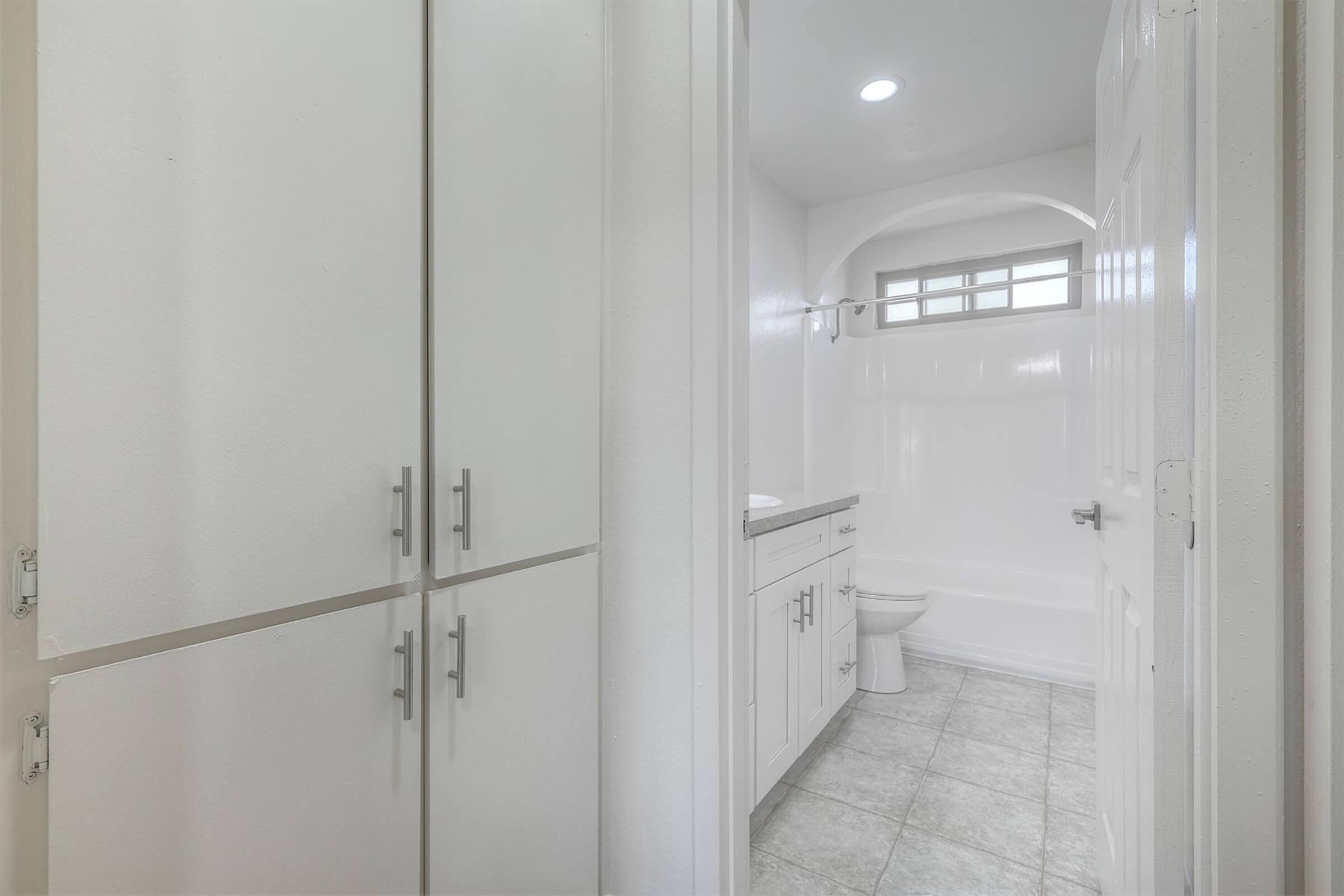 A clean, bright bathroom featuring white cabinetry, a bathtub, and a toilet. The space is modern with a tiled floor and a small window providing natural light. The walls are painted in a light color, enhancing the spacious feel.