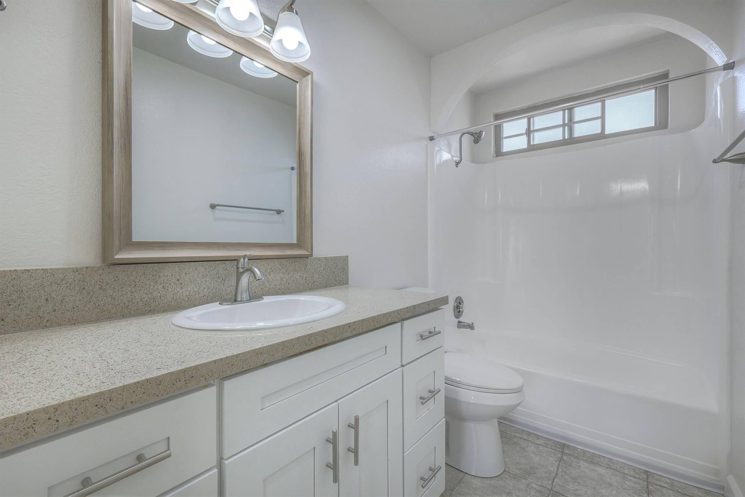Modern bathroom featuring a white vanity with a sink, a large mirror, and three light fixtures above. The bathroom includes a bathtub with a shower, a toilet, and light-colored tile flooring. The walls are painted white, creating a bright and clean atmosphere.