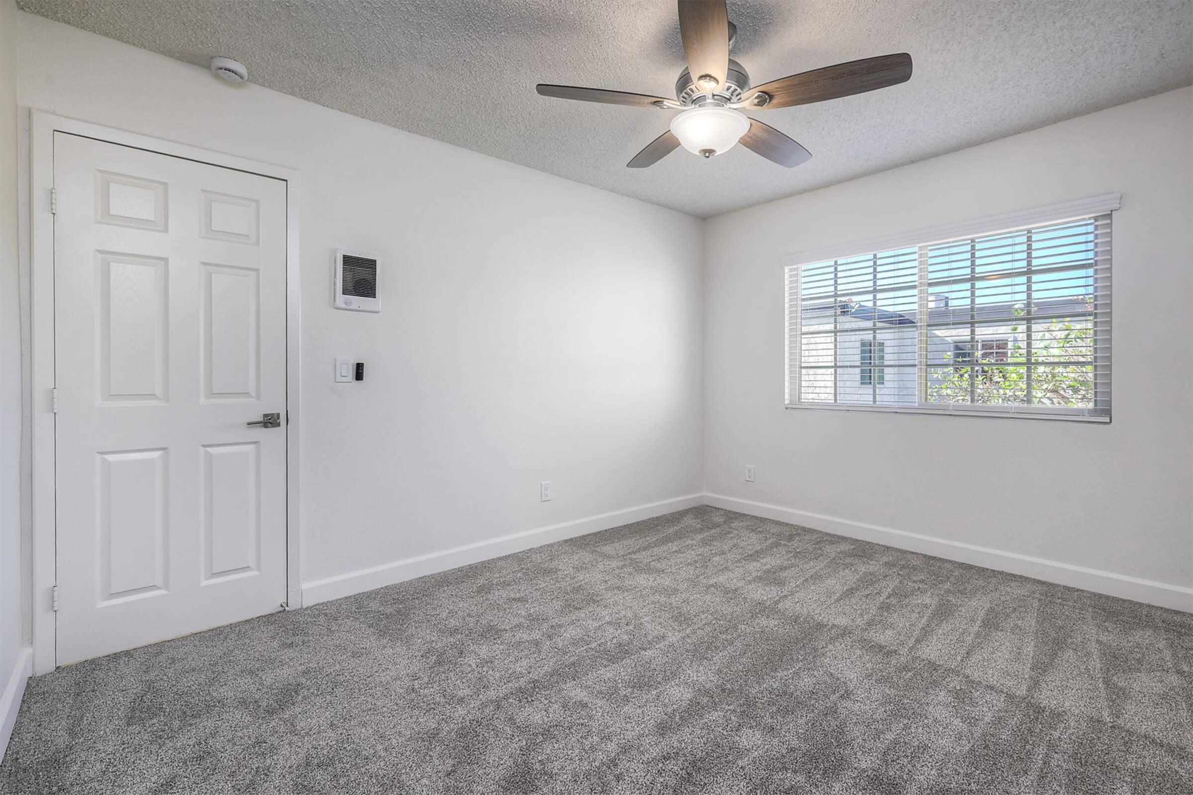 Empty room with light gray carpet, white walls, and a ceiling fan. A door on the left leads to the hallway, while a window on the right allows natural light to enter. The window has white blinds and views of the outside. A small wall-mounted unit is visible near the door.