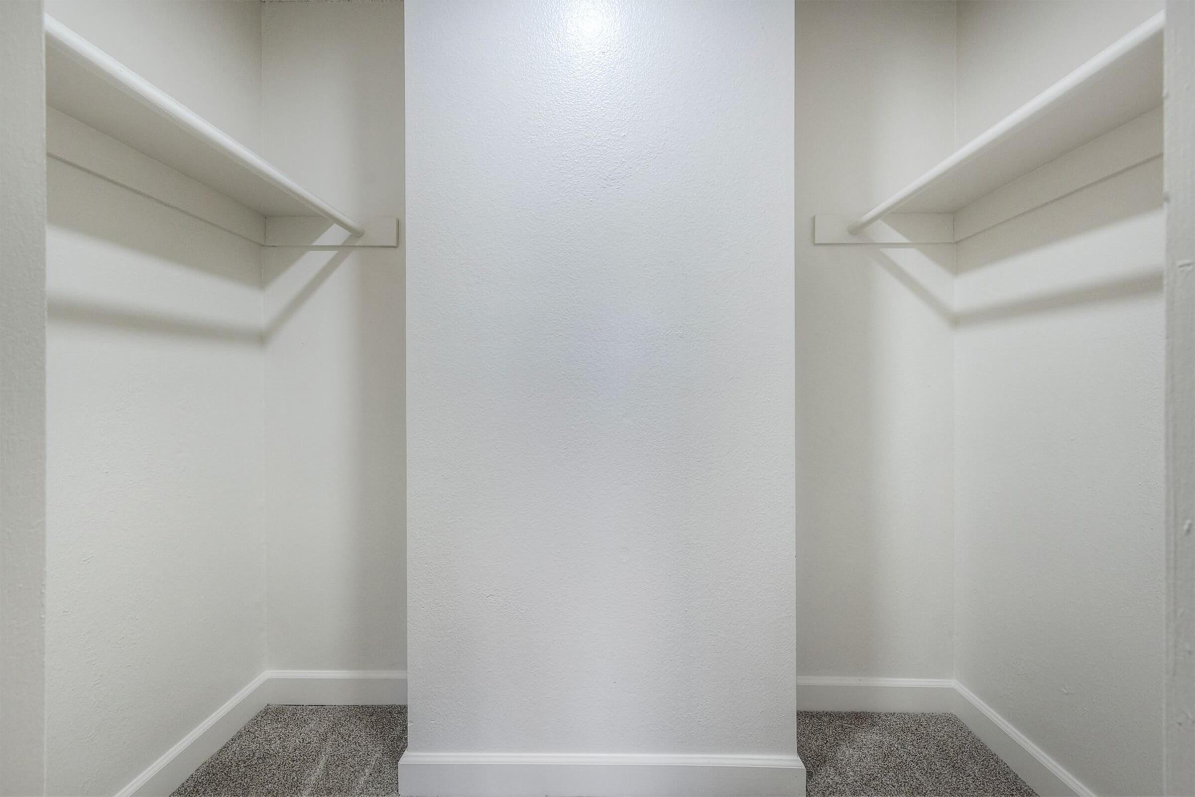 A blank, empty closet with white walls and two shelves on either side. The floor is carpeted, and the space is well-lit, creating a clean and minimalistic appearance.