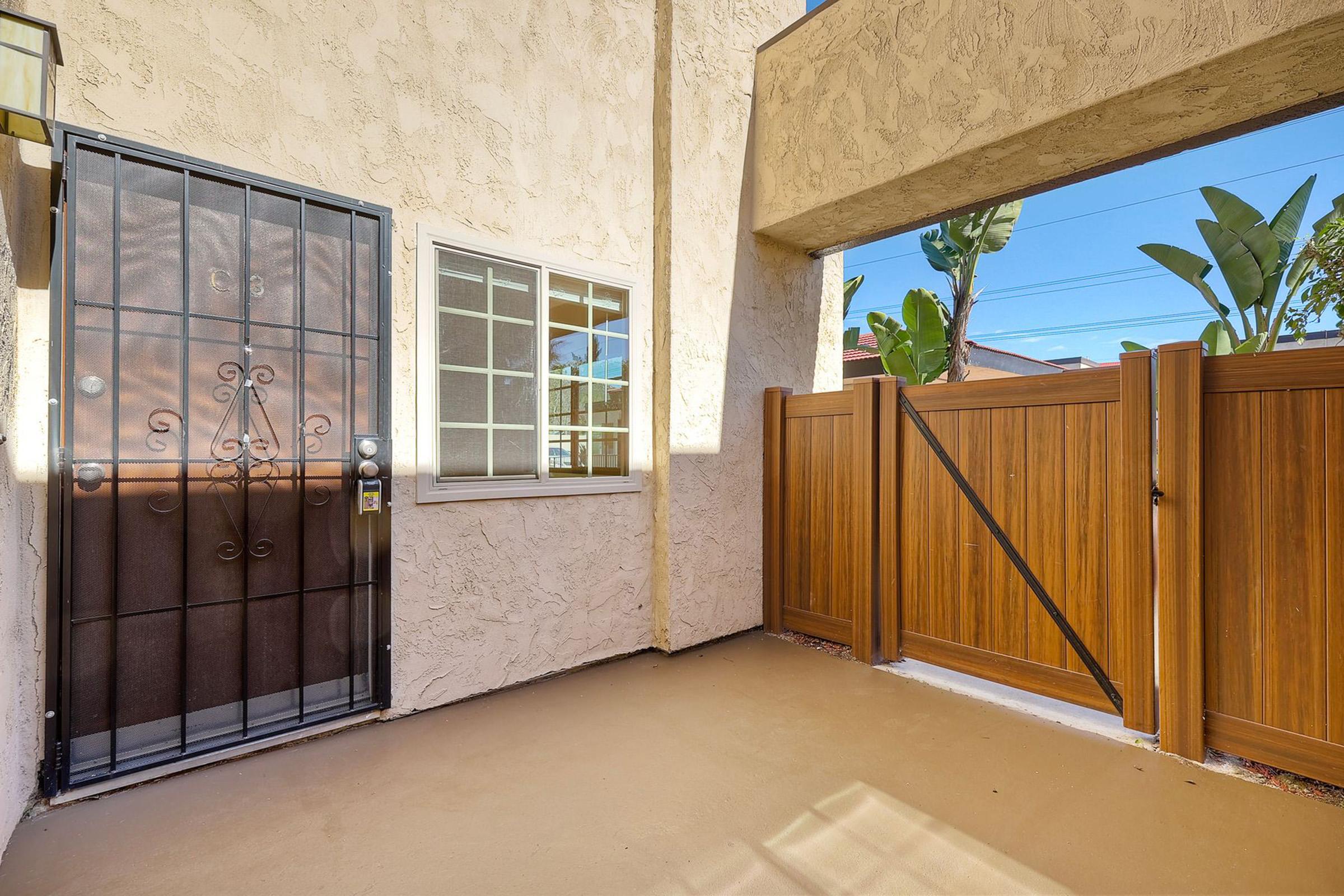 A small outdoor area featuring a gated entrance with a decorative metal door, a window beside it, and a wooden fence. Surrounding the area are tall green plants. The ground is a light color, and the space is well-lit by sunlight.
