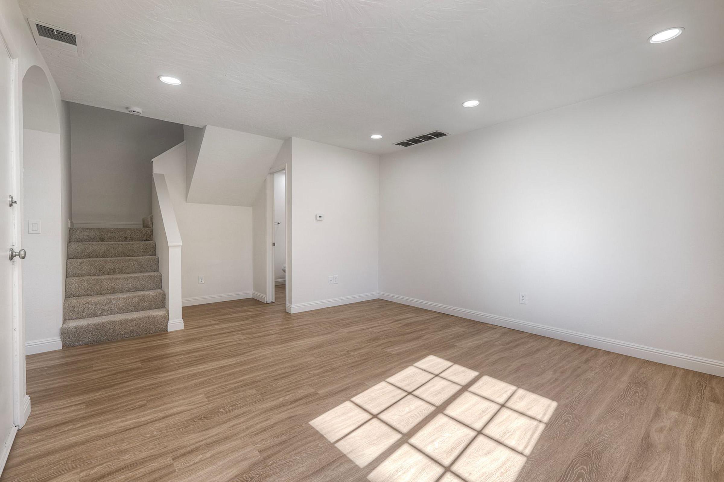 A simple, empty room featuring light-colored walls and hardwood flooring. Natural light enters through a window, casting a shadow on the floor. Stairs lead to an upper level, and there are recessed lighting fixtures on the ceiling. The space appears bright and airy, suitable for various uses.