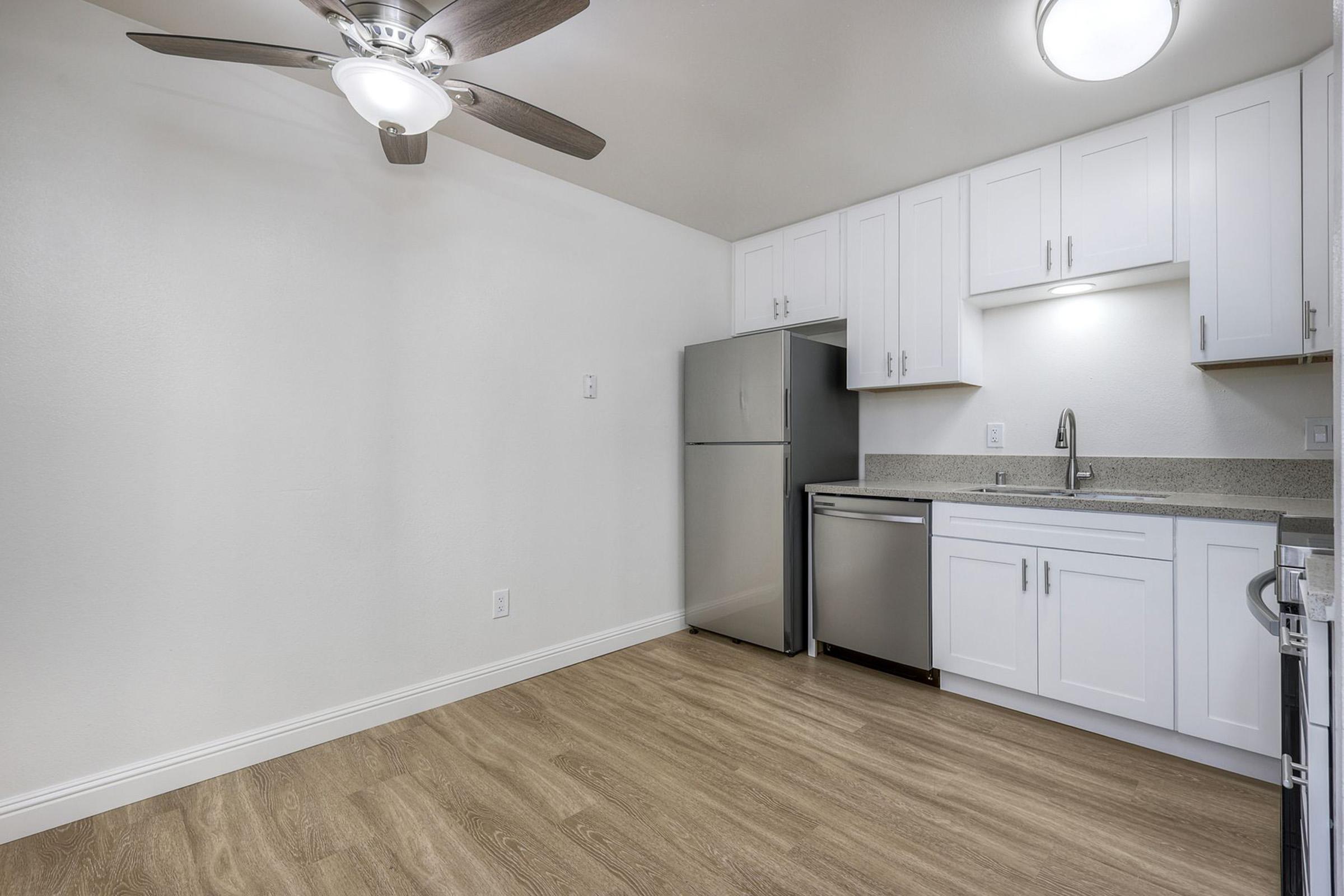 A modern kitchen featuring a stainless steel refrigerator, a dishwasher, and a sink with a sleek countertop. The room has white cabinets, a ceiling fan, and light-colored wooden flooring, creating a bright and airy atmosphere. The walls are painted white, giving it a clean and contemporary look.