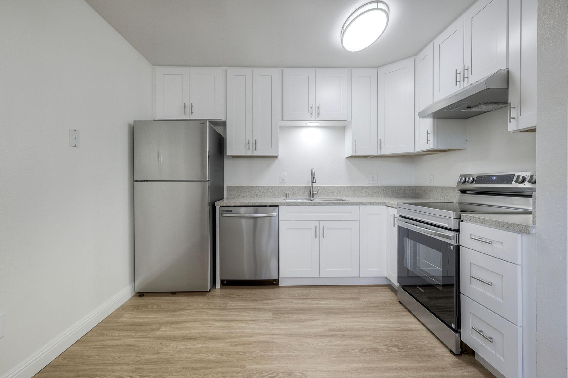 Modern kitchen with stainless steel appliances including a refrigerator, dishwasher, and oven, white cabinetry, and grey countertops. Bright lighting from a ceiling fixture, light wood flooring, and white walls create an airy atmosphere.