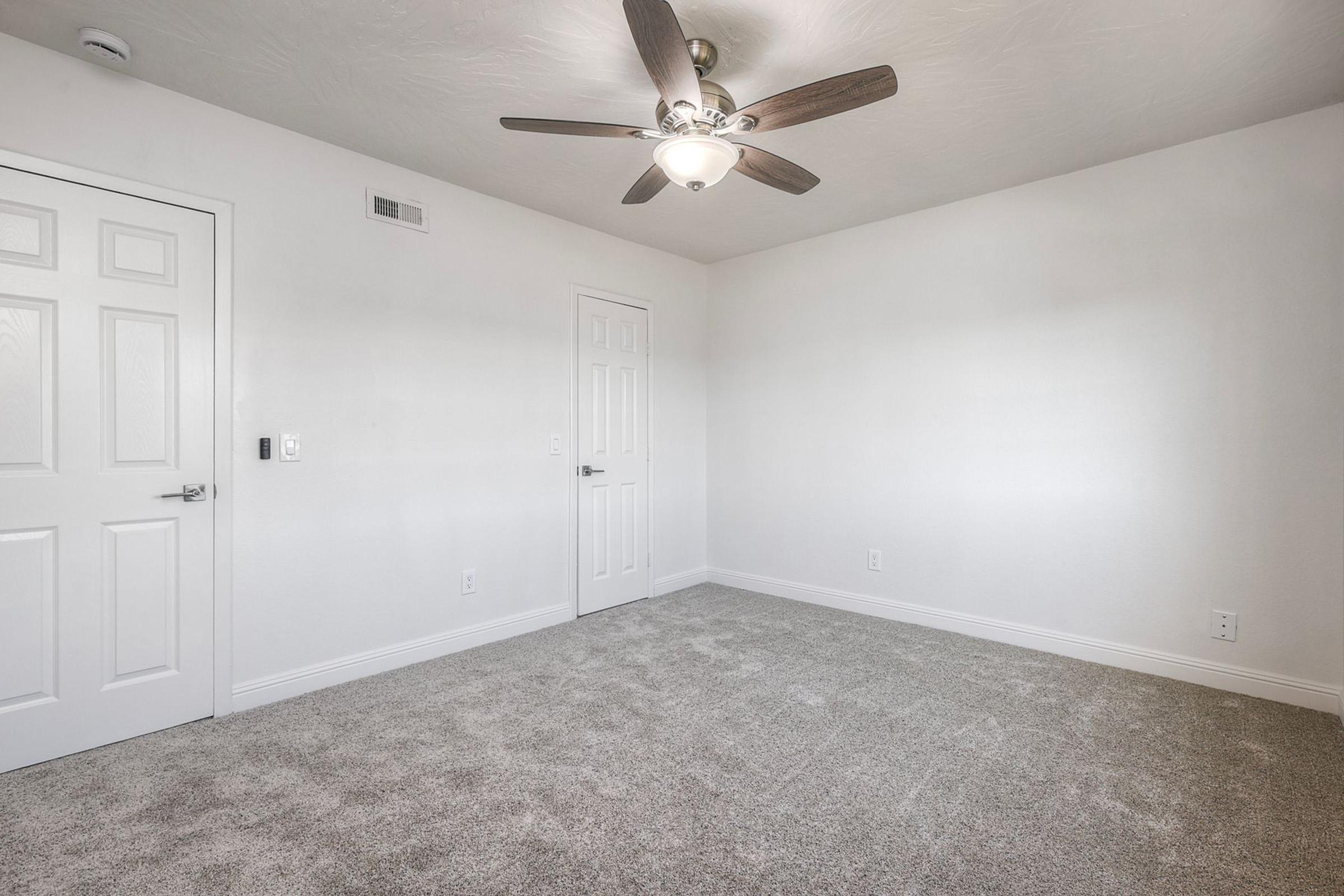 A well-lit, empty room featuring light gray carpet and neutral-colored walls. The ceiling has a modern ceiling fan, and there are two closed doors, one leading to a closet. The room is spacious and unfurnished, creating a blank canvas for potential decoration.