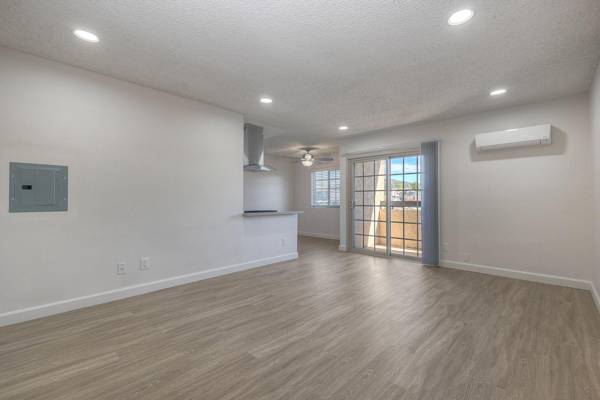 A spacious, empty living area featuring light-colored walls and wood-style flooring. It includes a sliding glass door leading to a balcony, a ceiling fan, and a kitchen area with a countertop. Natural light fills the room, creating an inviting atmosphere.