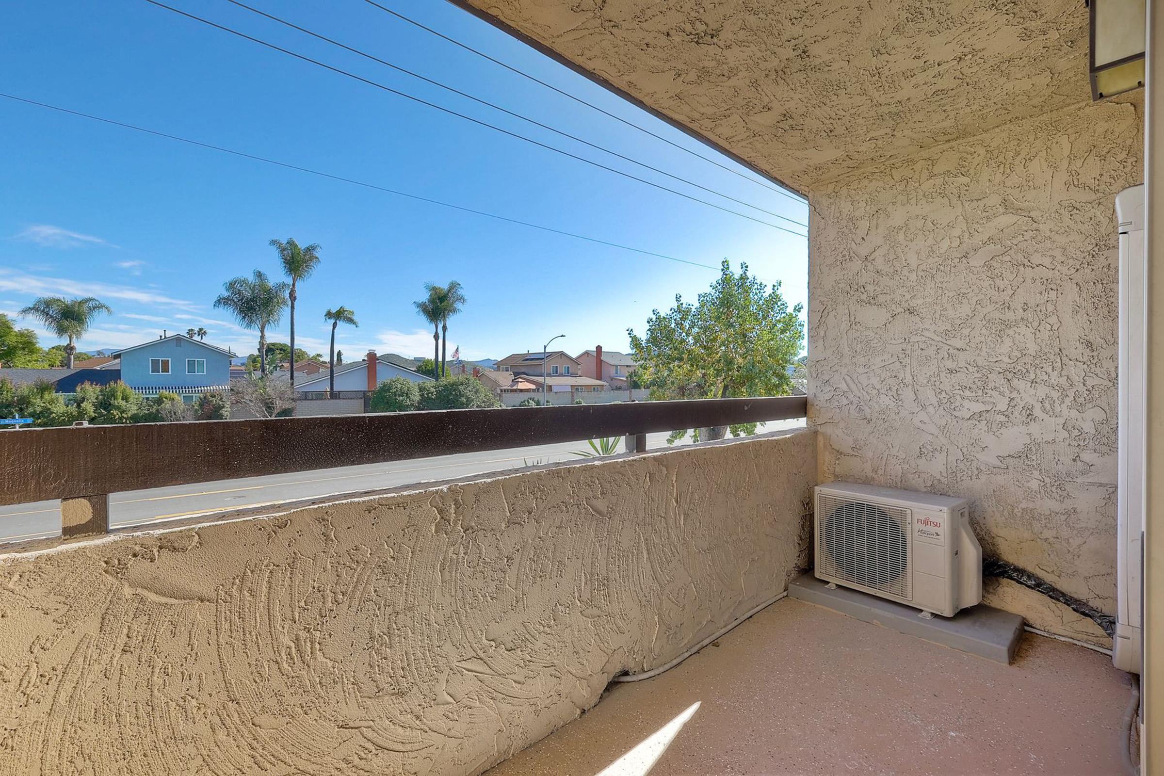 A balcony view featuring a clear blue sky, palm trees, and residential houses in the background. An air conditioning unit is installed on the wall of the balcony. The surface of the balcony is plain and textured, with sunlight illuminating the scene.