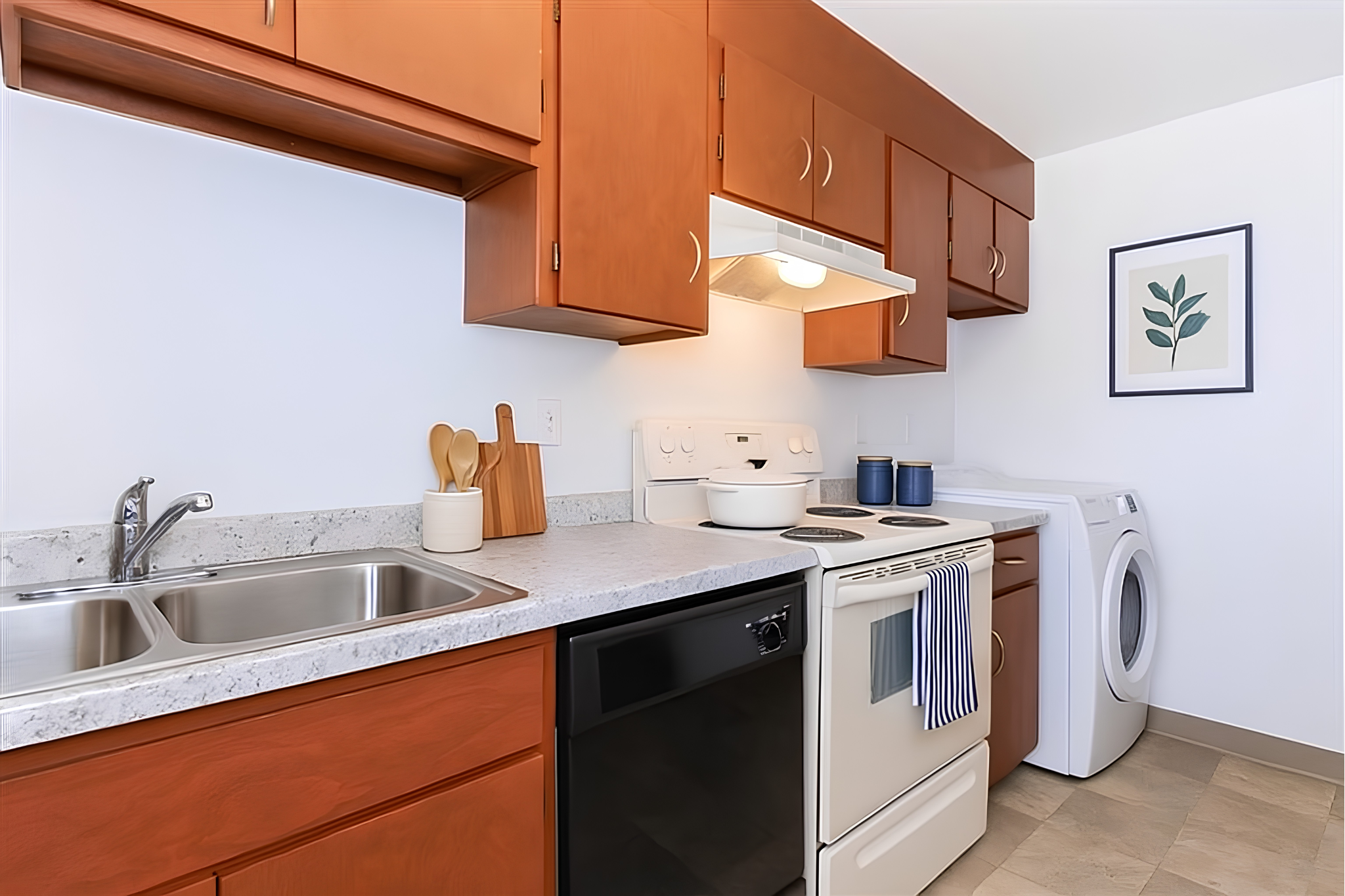 A modern kitchen featuring wooden cabinets, a double sink, a stove, and a dishwasher. Adjacent to the kitchen is a laundry area with a washing machine. The countertop is light-colored, and there are kitchen utensils displayed. A framed picture hangs on the wall, adding a decorative touch to the room.