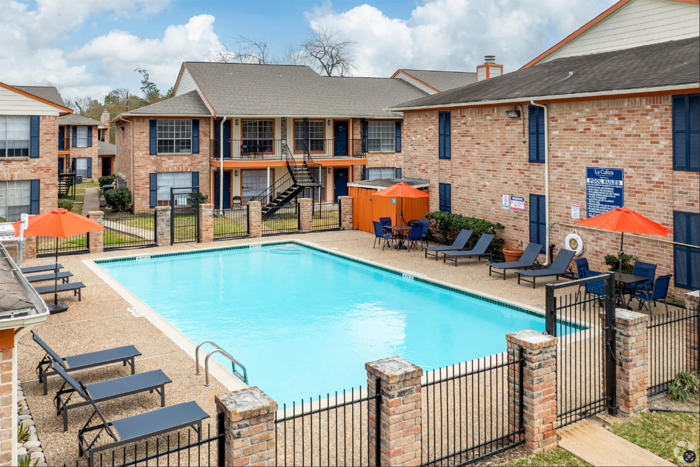 A well-maintained outdoor swimming pool surrounded by lounge chairs and orange umbrellas, located in an apartment complex. Brick buildings with balconies are visible in the background, under a partly cloudy sky. The pool area is enclosed with a black fence.