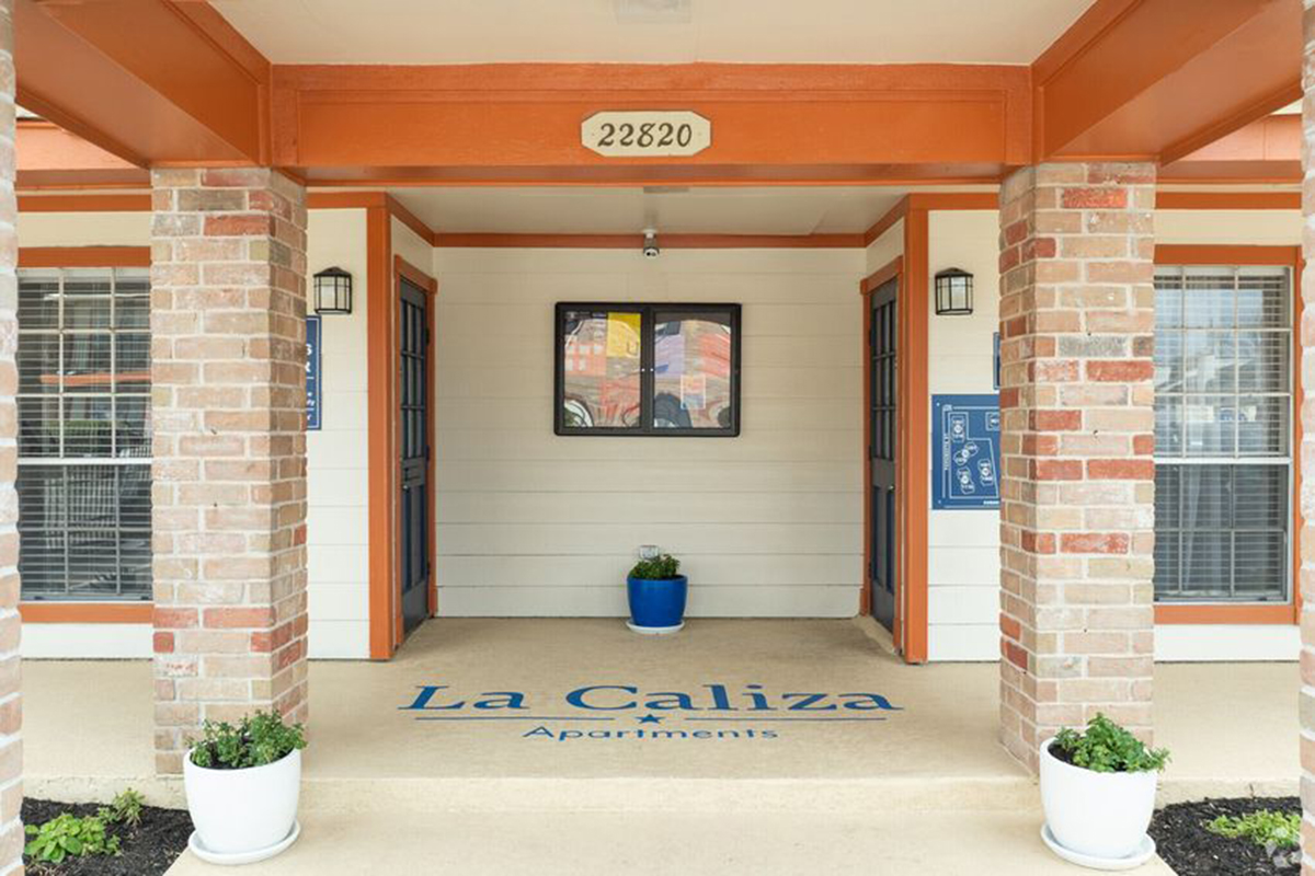 Entrance to La Caliza Apartments featuring a covered porch. The space includes brick columns, decorative wall lights, a potted plant in a blue pot, and a sign indicating the apartment name and number "22820" on the wall. There are two planters with greenery flanking the entrance.