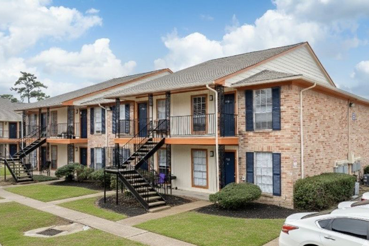 Two-story brick apartment building with orange accents and black railings, featuring a landscaped lawn with shrubs. The building has multiple units with balconies and stairs leading to each entrance, set against a partly cloudy sky. A parked white car is visible in the foreground.