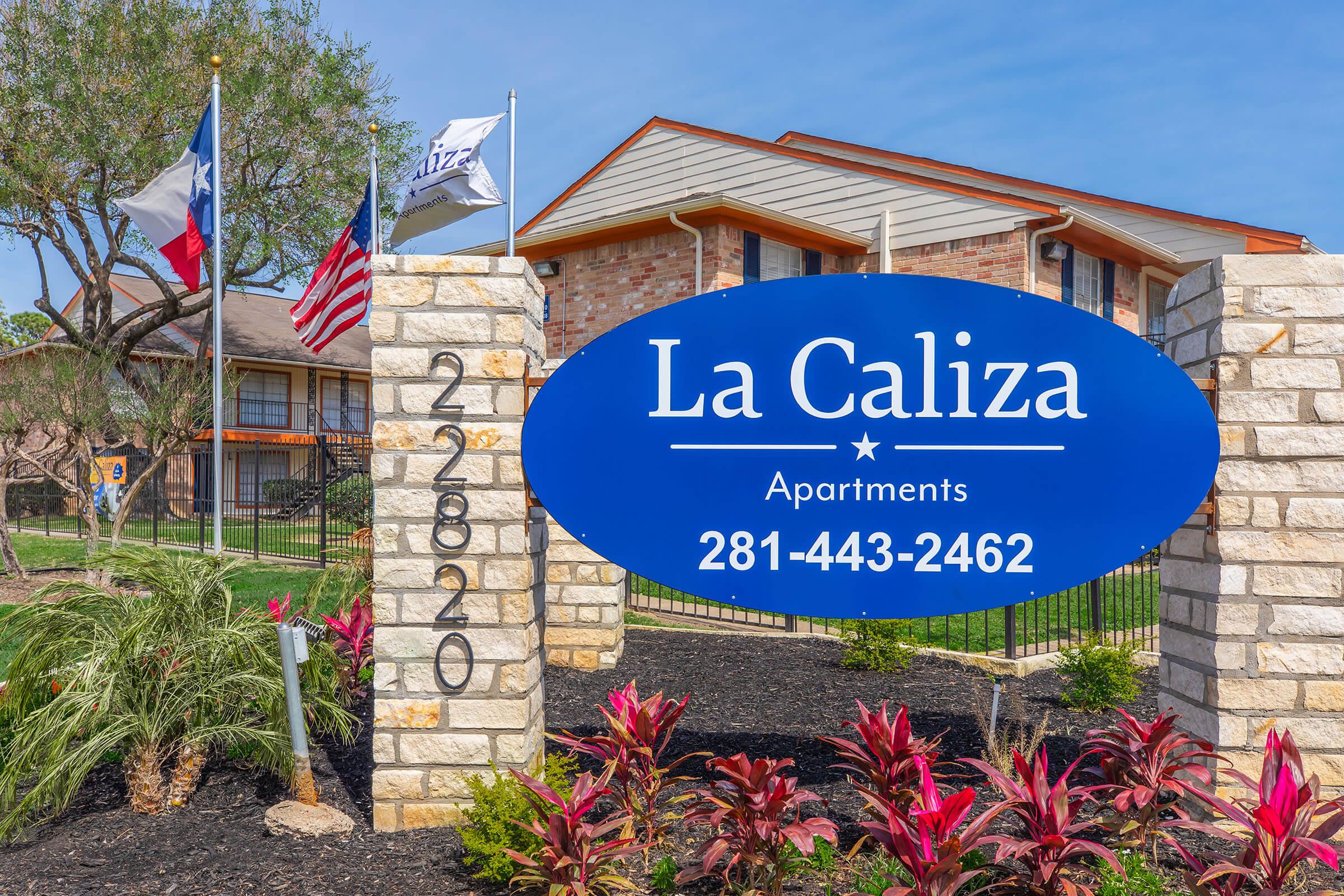 Sign for La Caliza Apartments displaying the name in white text on a blue background, along with the contact number 281-443-2462. Flags of Texas and the United States are visible, with a landscaped area featuring plants and shrubs in front of the stone pillars supporting the sign.