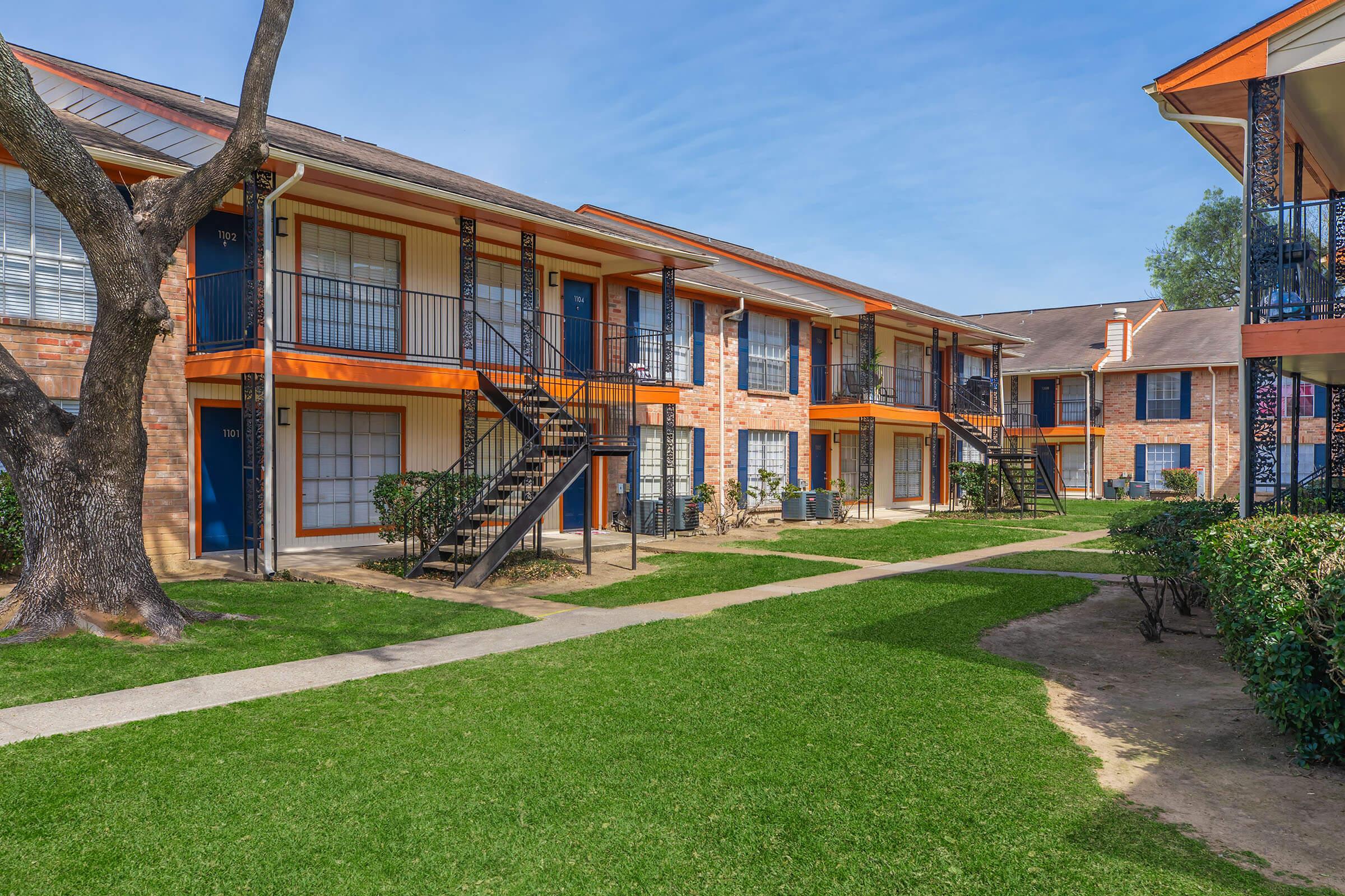 A sunny view of an apartment complex featuring two-story buildings with orange accents and black railings. Lush green grass and well-maintained pathways separate the units, and trees are present in the foreground, creating a welcoming community atmosphere.