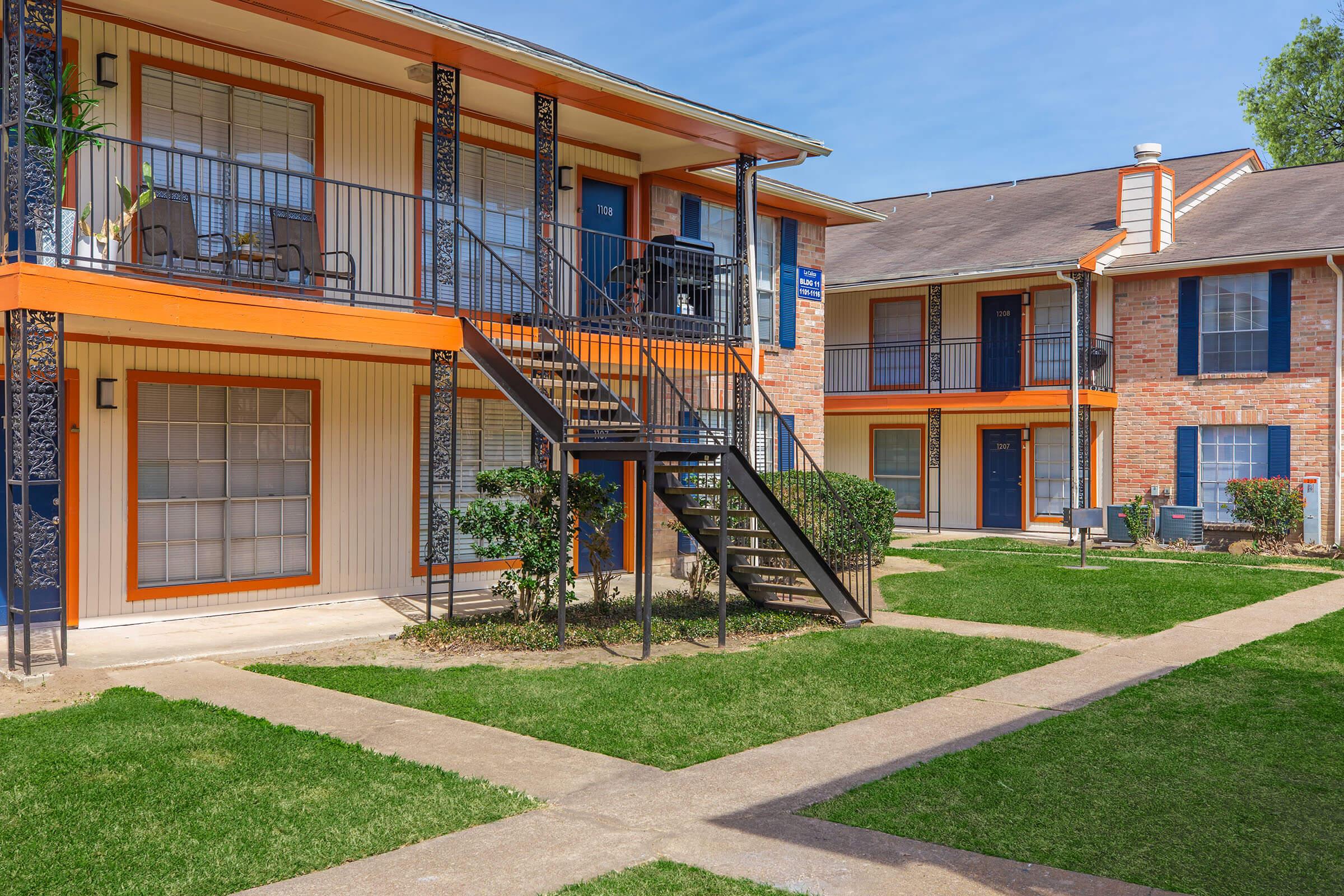 Two-story apartment building with orange accents and a staircase leading to the second floor. Green lawns surround the building, with concrete pathways connecting the entrances. Windows are adorned with curtains, and there are small shrubs in front. Clear blue sky above.