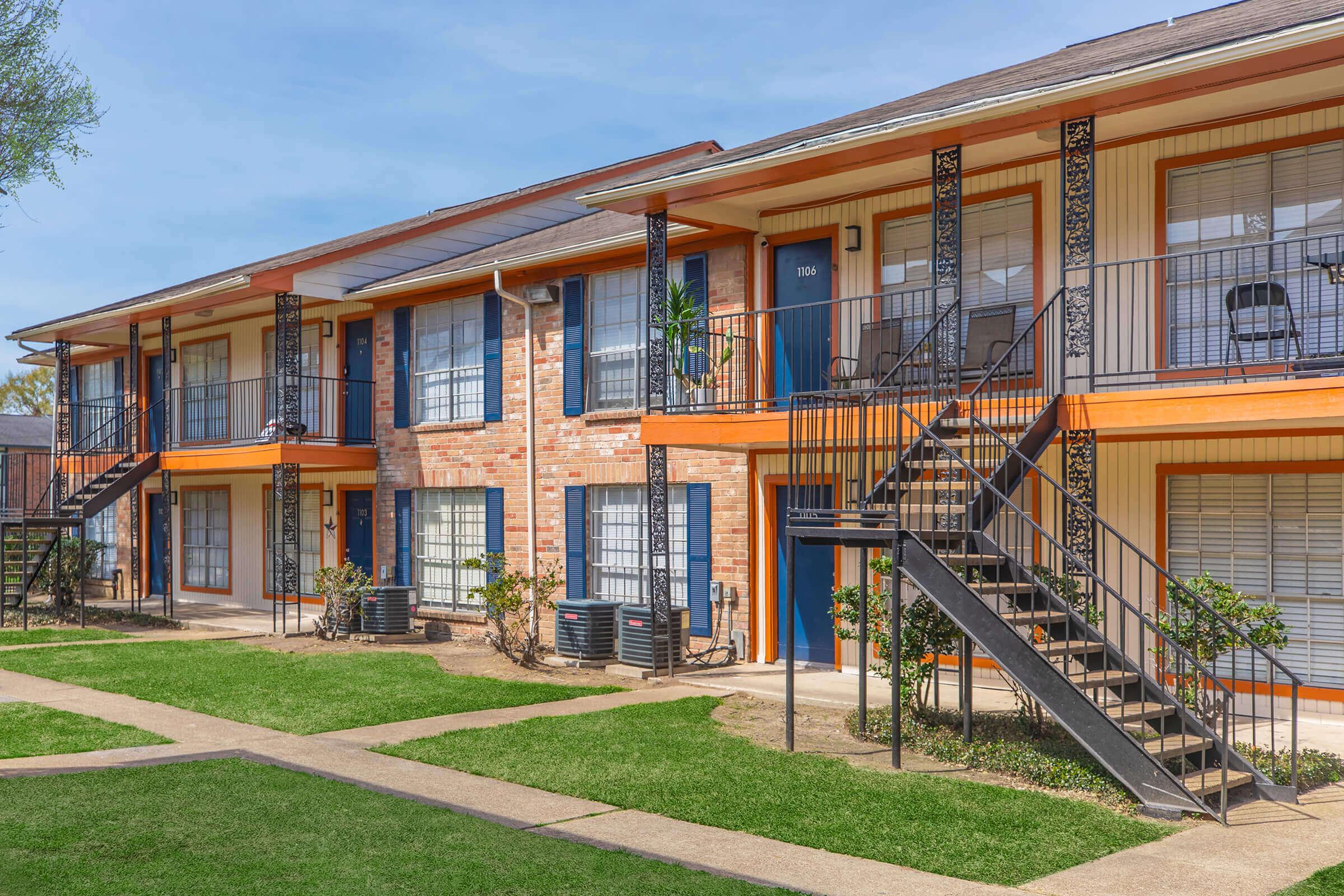 A two-story apartment building featuring a brick facade with orange trim. Staircases lead to the upper level, and there are multiple doors with windows on each floor. The surrounding area has well-maintained green grass and pathway. The sky is clear and blue, indicating a sunny day.