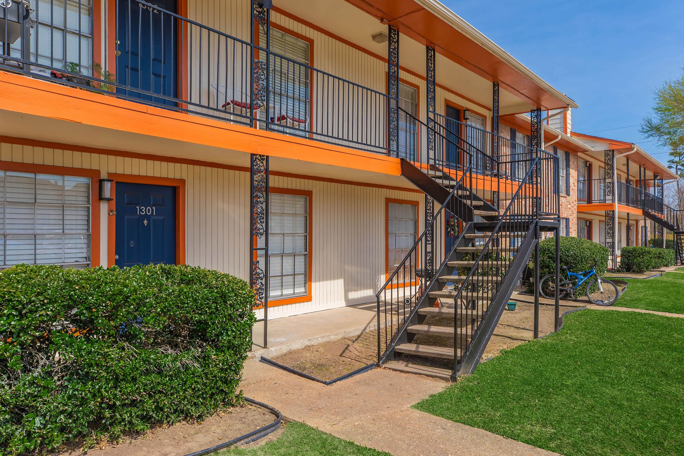 A view of an apartment building with orange accents, featuring two levels of balconies. A black metal staircase leads to the upper level, and green grass borders the pathway. A blue bicycle is parked beside the building, with a clear blue sky in the background.