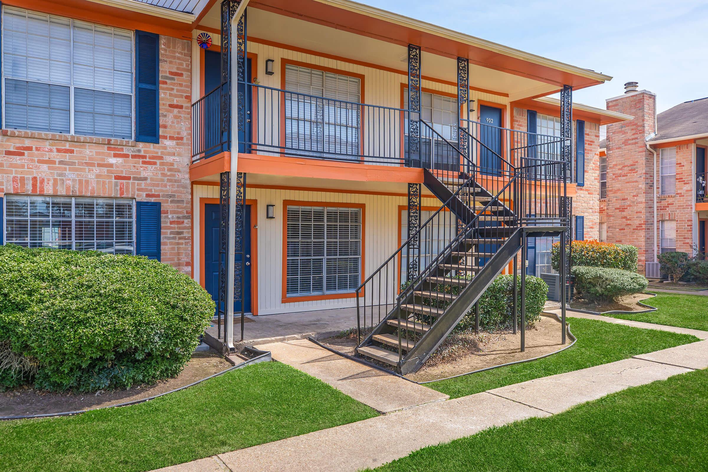A two-story apartment building with orange accents and black iron stair railings. Lush green shrubs are near the entrance, and well-maintained lawns lead to a concrete pathway. The windows have white blinds, and the building features red brick exteriors with blue shutters.