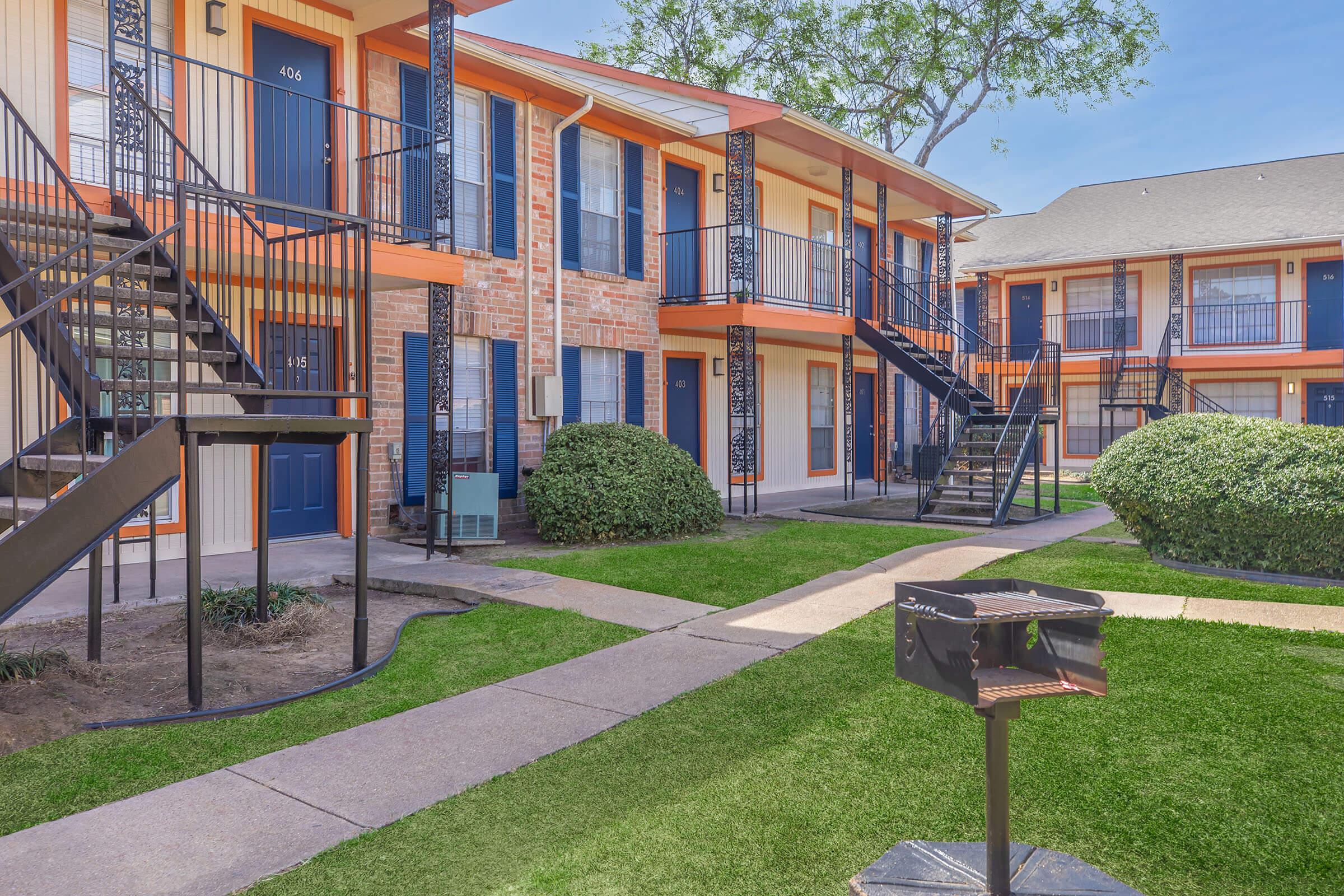 A view of a multi-unit apartment complex with two buildings, featuring orange accents, black railings, and staircases. The well-manicured lawn includes a BBQ grill near the walkway and small bushes surrounding the area. Clear blue skies complement the scene, creating a welcoming atmosphere.
