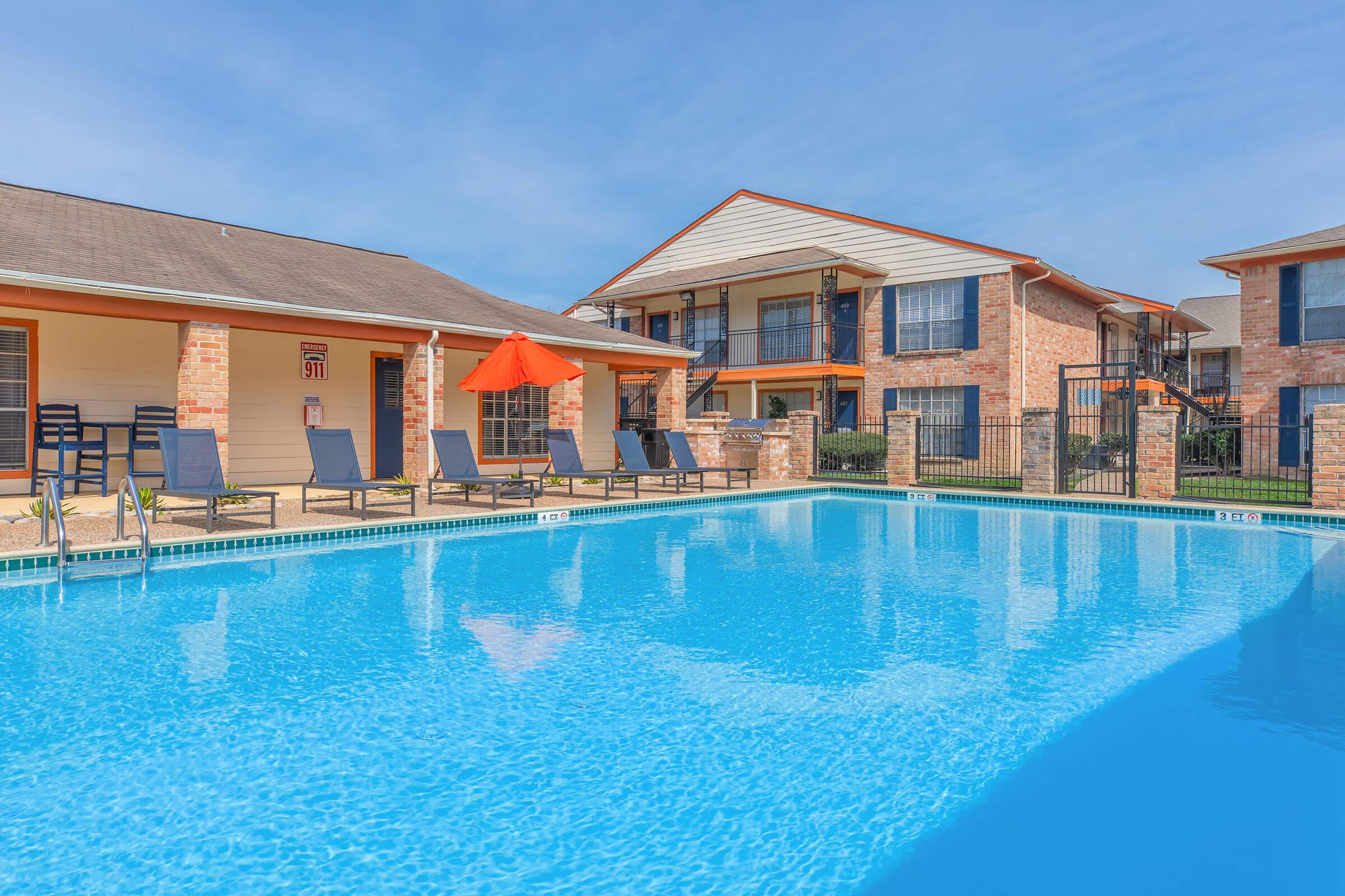 A clear blue swimming pool surrounded by lounge chairs and an orange umbrella. Adjacent to the pool are brick buildings with balconies, under a blue sky. The setting suggests a relaxed, inviting atmosphere ideal for leisure and sunbathing.