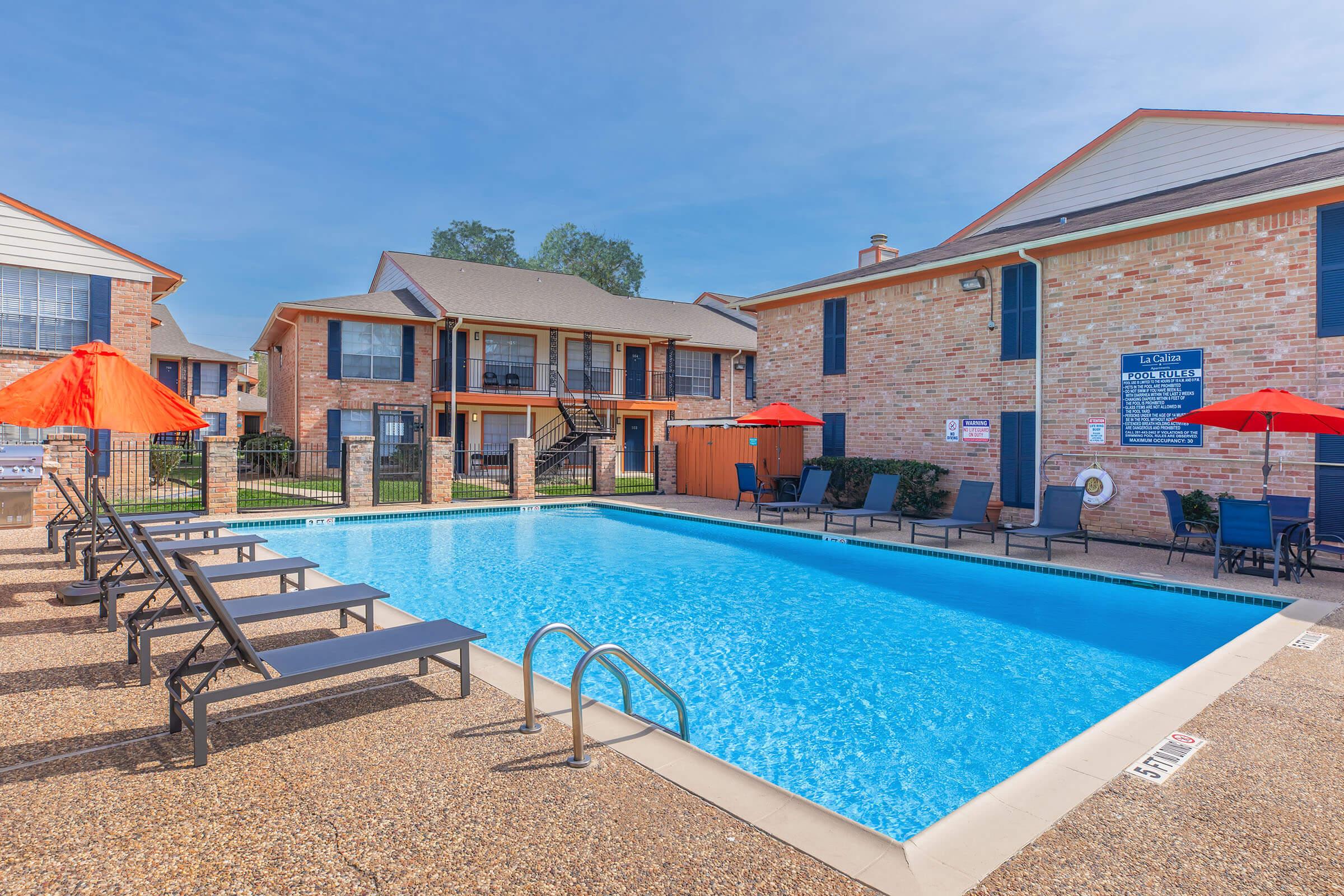 A clear blue swimming pool surrounded by lounge chairs and red umbrellas, with brick buildings in the background. The area has well-maintained landscaping and a sunny sky, creating a relaxing atmosphere.