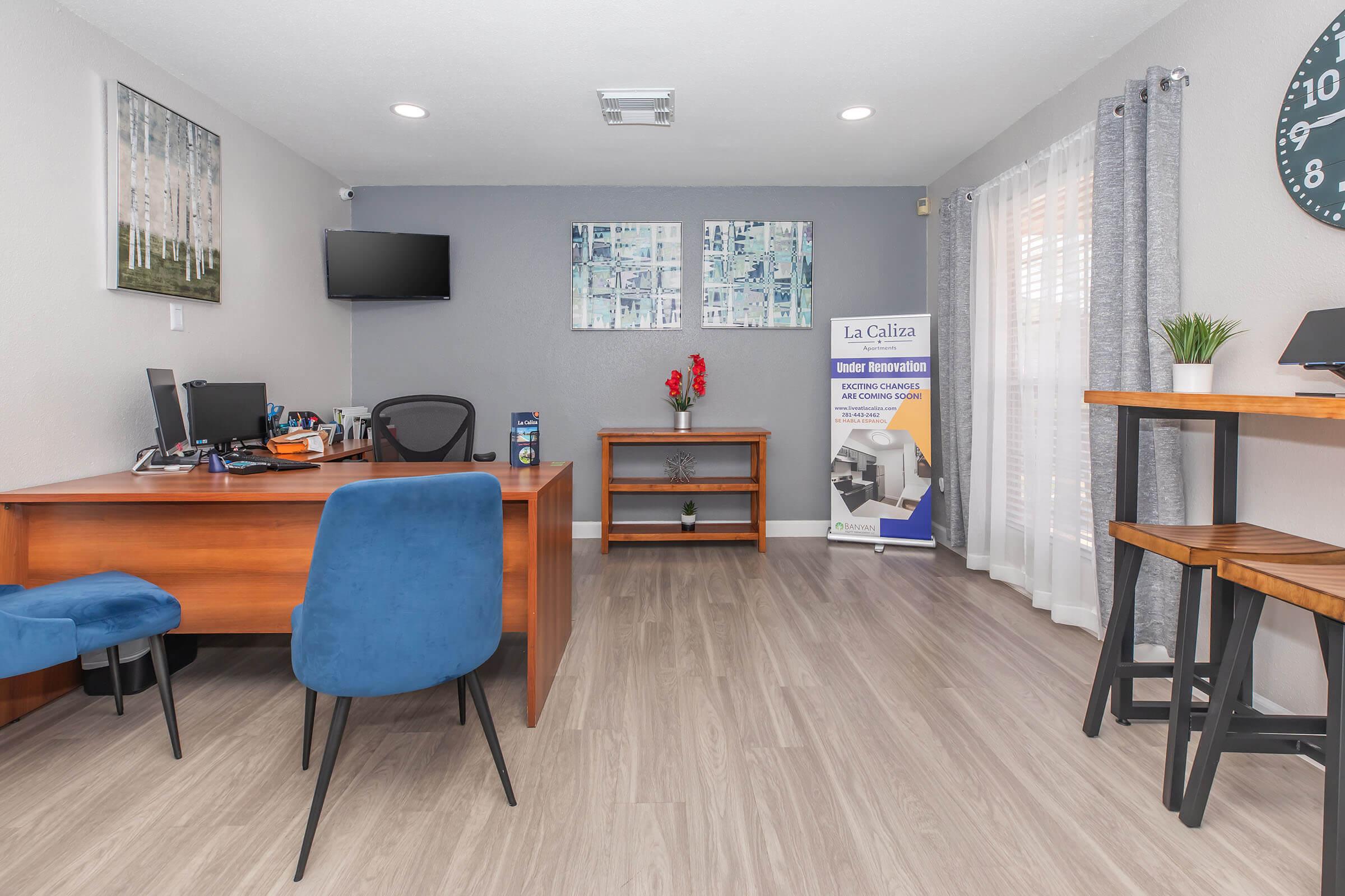 A modern office space with wooden furniture, including a desk and shelves. A computer setup is on the desk, with a black chair and blue accent chairs. A wall clock and decorative artwork adorn the walls. A banner for "La Caliza" is visible, promoting services, alongside natural light from windows.